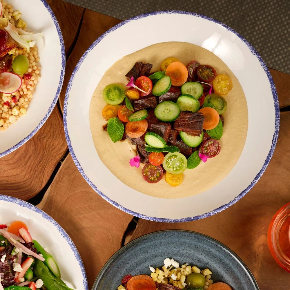 Overhead view of a white plate spread with house chickpea hummus topped with seared beef tenderloin tips and heirloom cherry tomato salad from the Mezza slip in Farewinds Market Hall.