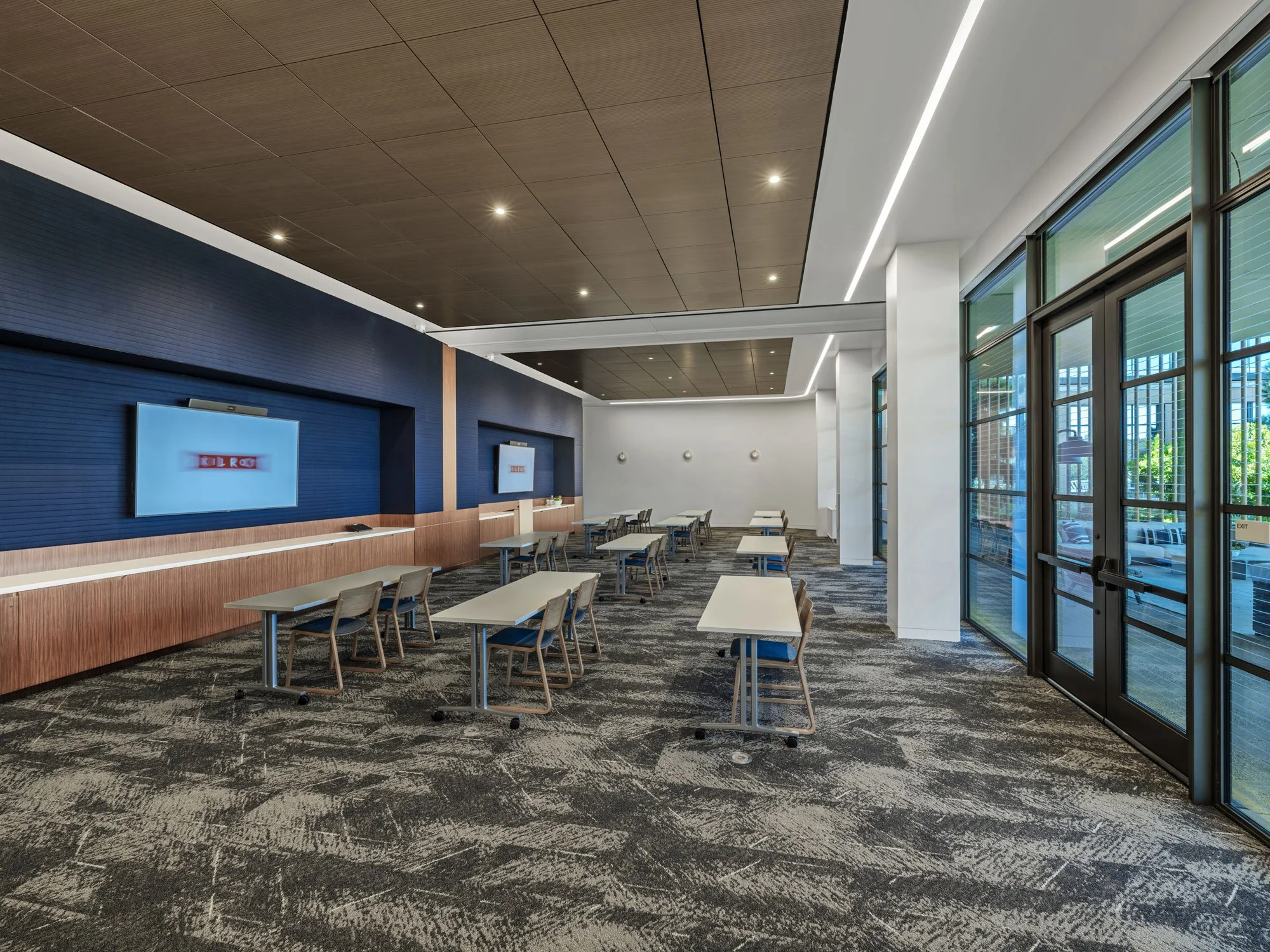 A conference room at the Farewinds Conference Center with rows of small white tables with two chairs, two 85-inch TV monitors on the wall at the front of the room, and glass wall opeining on to a patio at the back of the room.