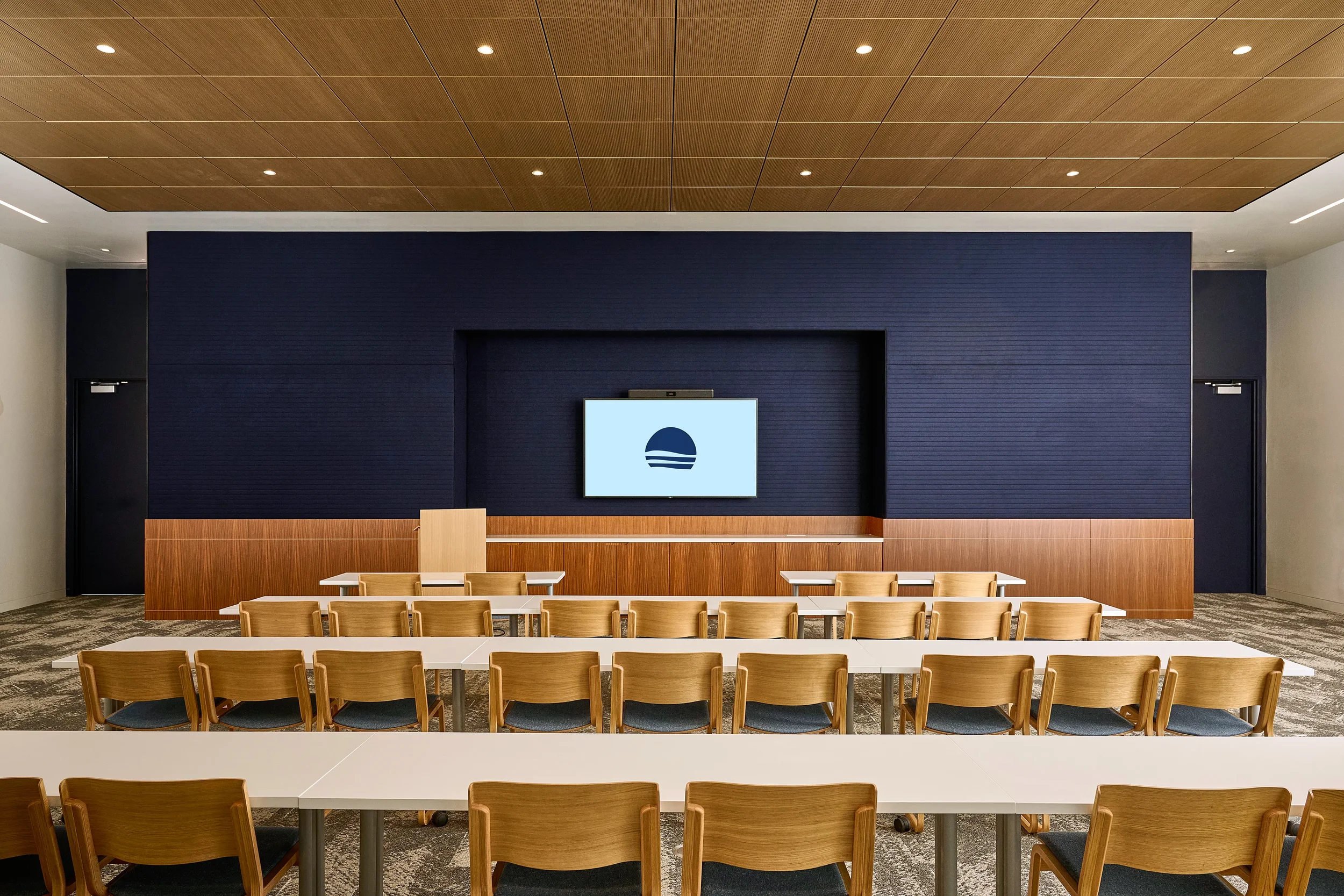 A conference room at the Farewinds Conference Center with long white tables, wooden chairs along one side of each table, and a 85-inch TV monitor on the wall at the front of the room.