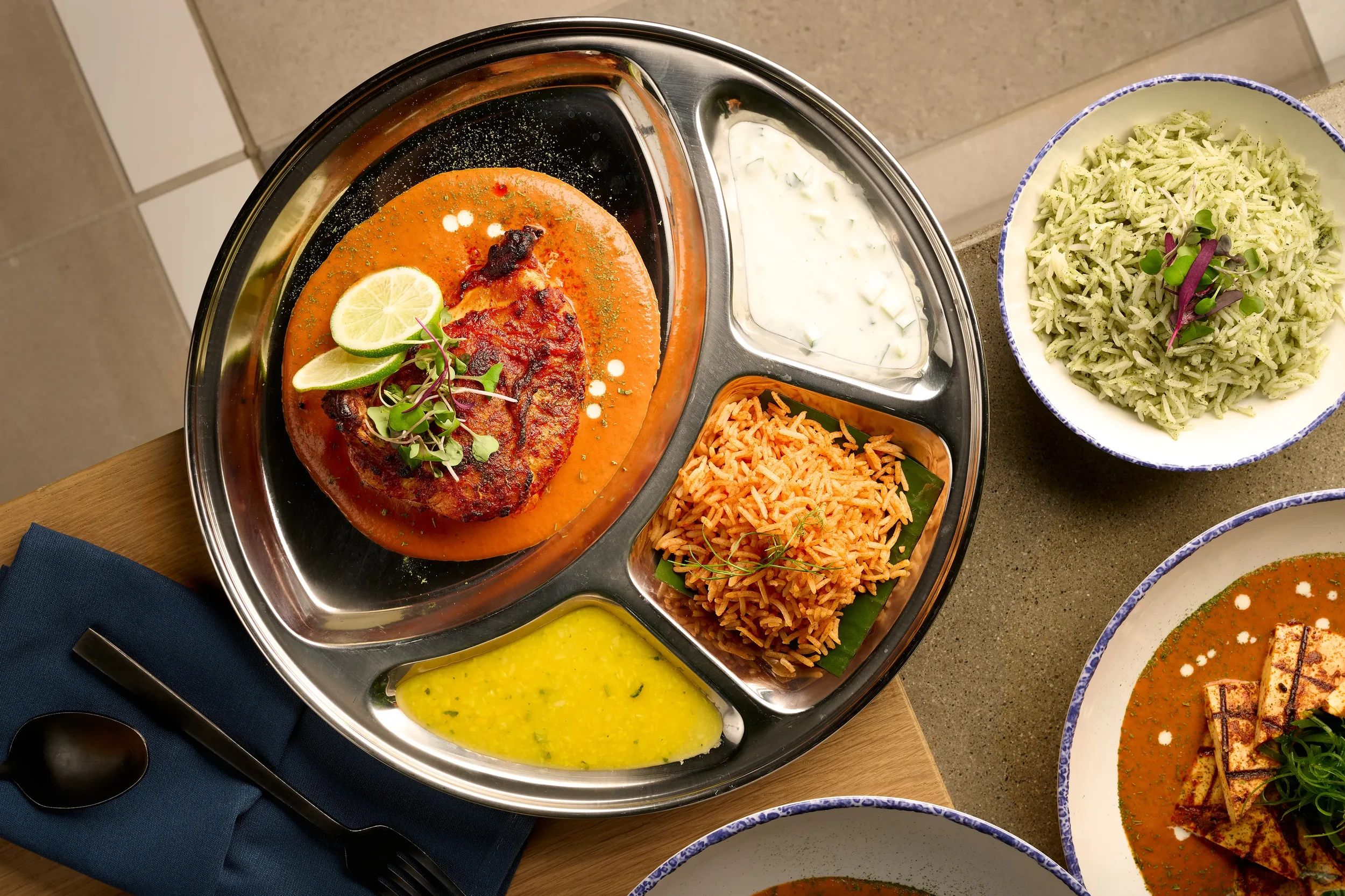 Overhead view of a thali plate with grilled chicken in smoked tomato yogurt sauce, yellow dal, rice, and raita from the Indian slip in Farewinds Food Hall.