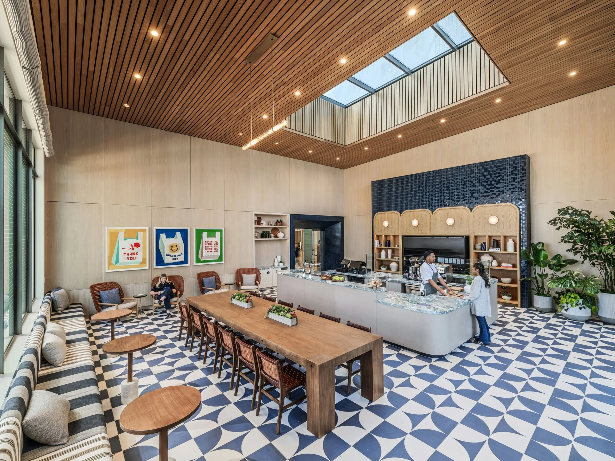 Reservable space in Farewinds Market with patterned tile floor, wooden ceiling, a long wooden table, booths and chairs along walls with round tables, and barista serving a customer.