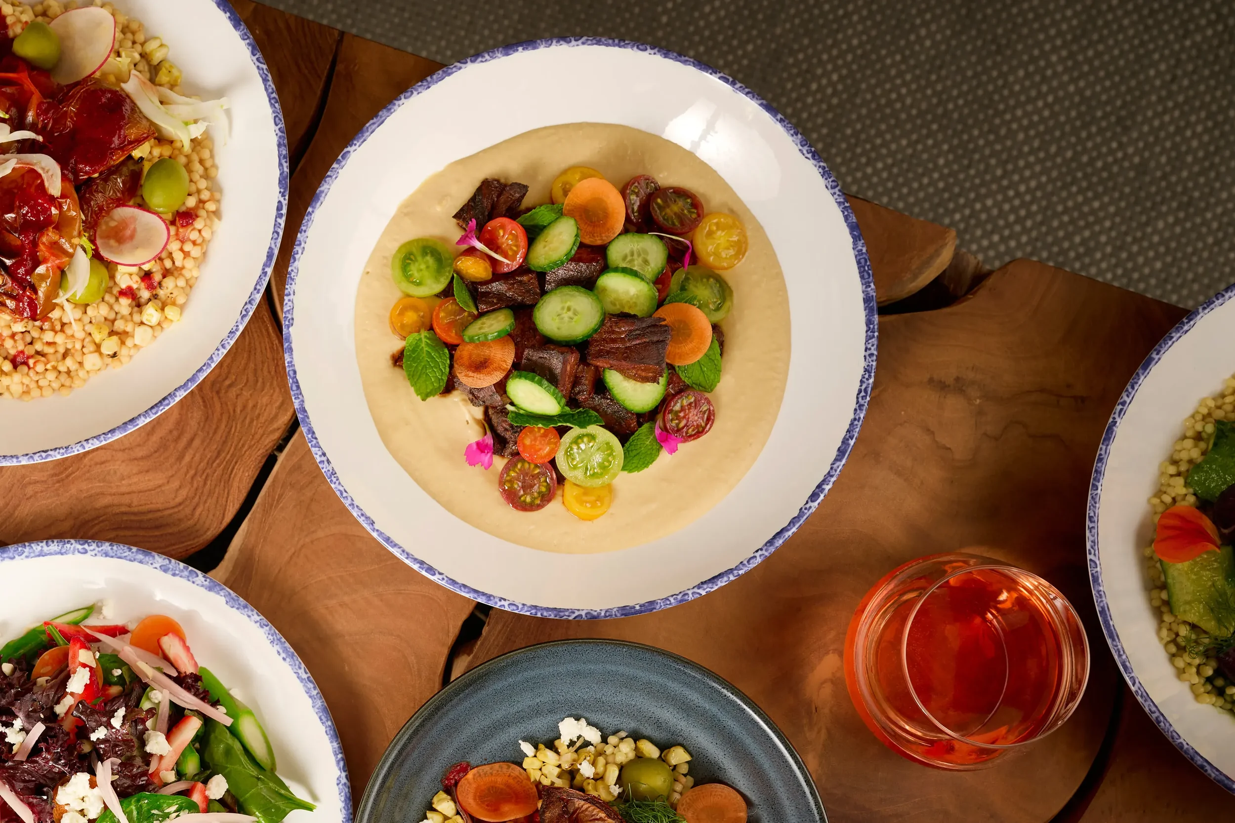 Overhead view of a white plate spread with house chickpea hummus topped with seared beef tenderloin tips and heirloom cherry tomato salad from the Mezza slip in Farewinds Food Hall.
