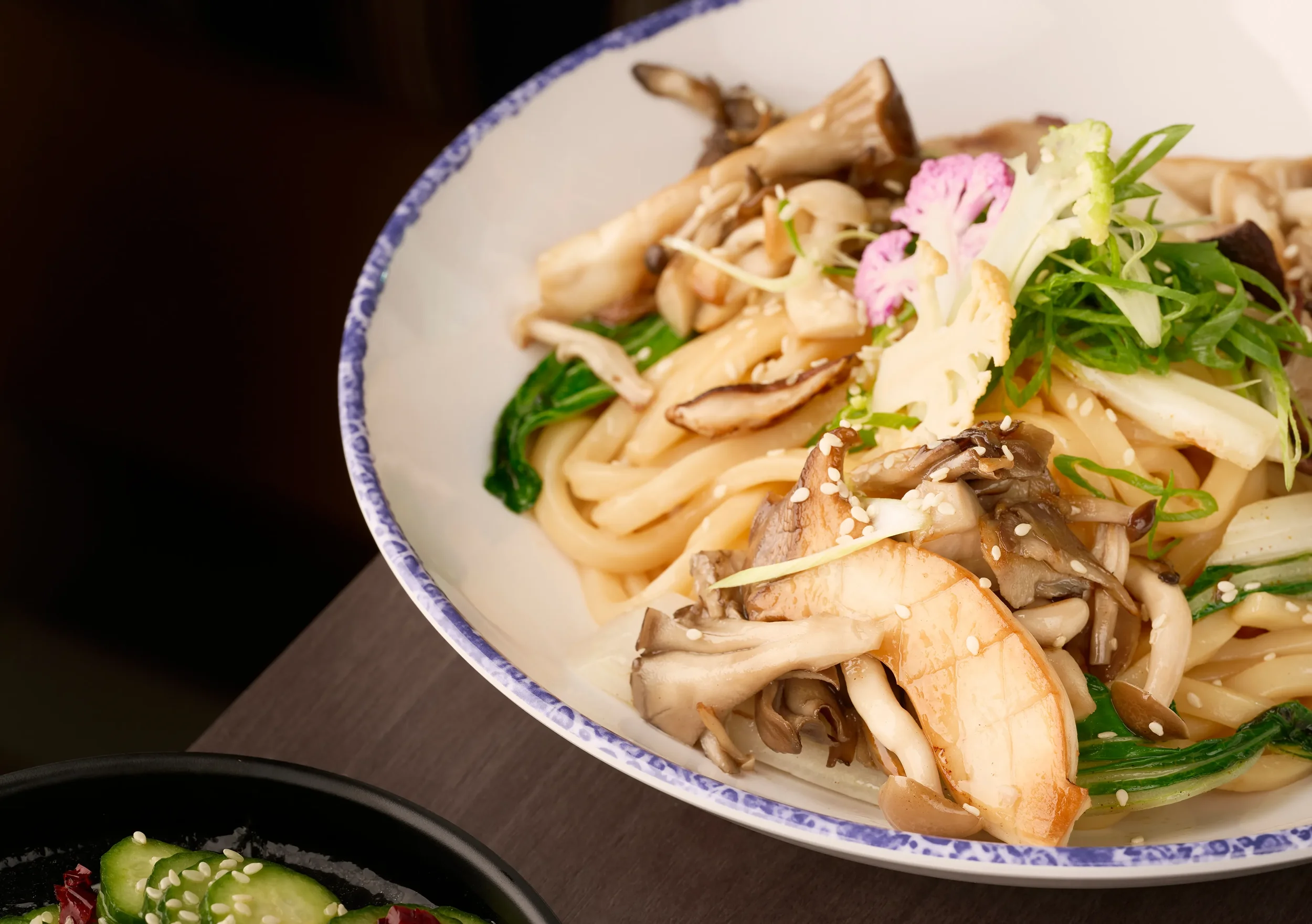 Close-up of a white bowl containing udon topped with wild mushrooms and bok choy from the Noodle Bar slip in Farewinds Food Hall.