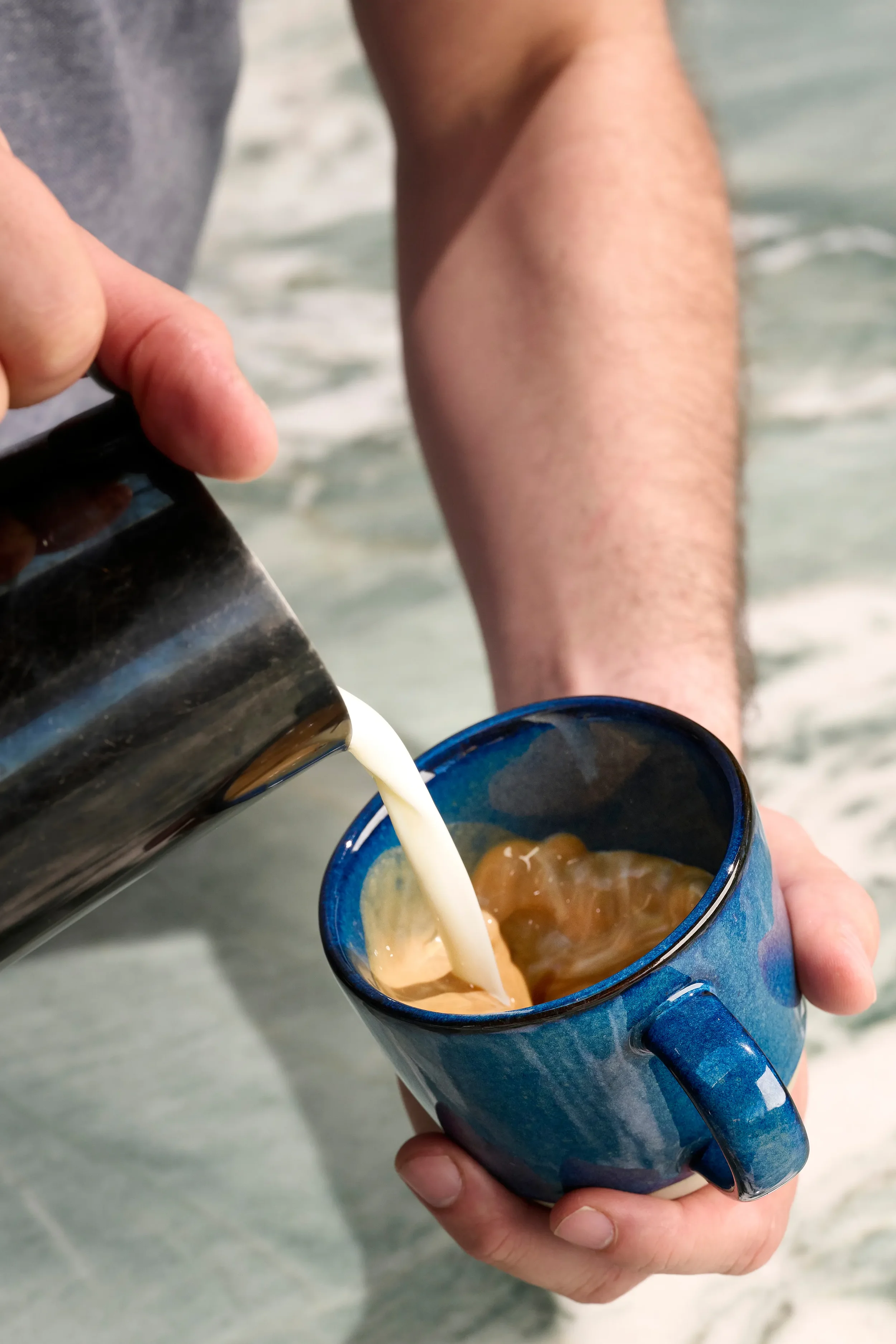Close-up of steamed milk being poured from a small metal pitcher into a blue ceramic cup with espresso at Farewinds Market.