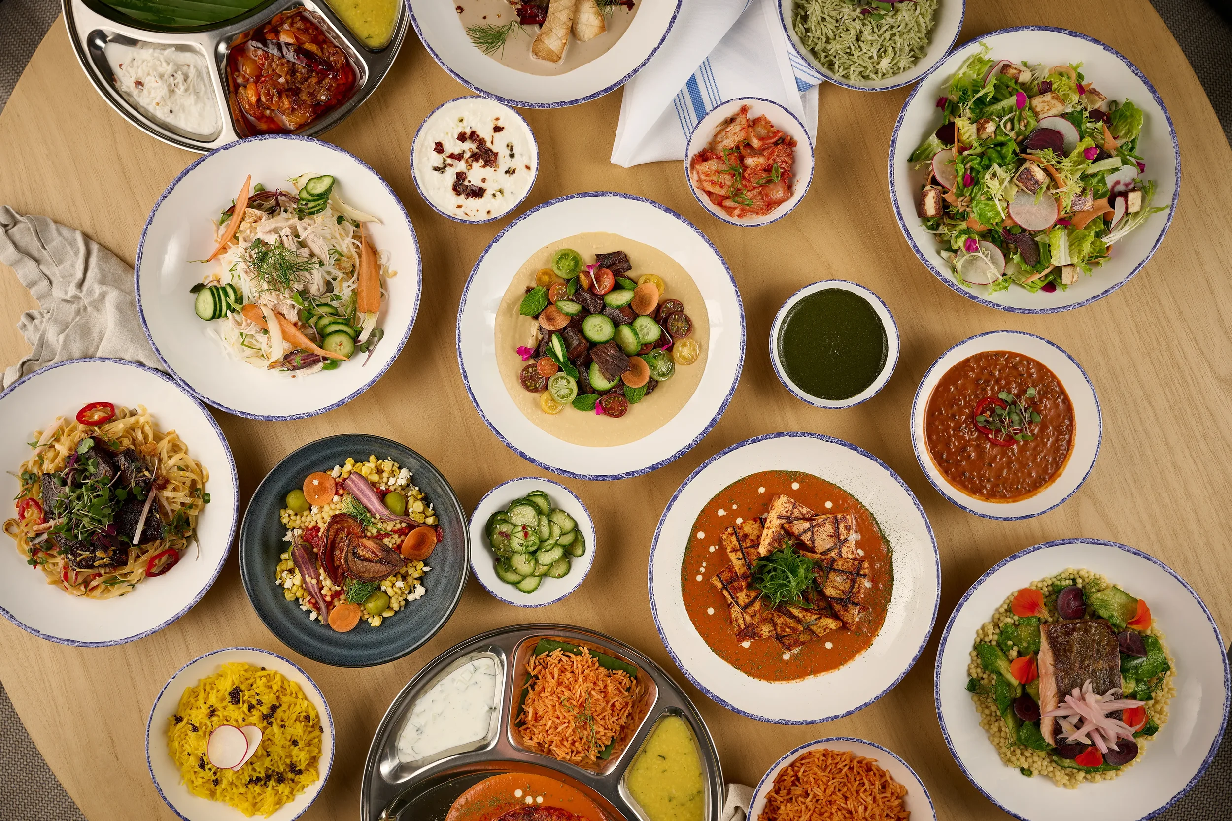Overhead view of various dishes from Farewinds Food Hall, including noodle bowls, ban chan, Indian curries with rice, salads, a hummus plate, and fish over couscous, arranged on a wooden table.