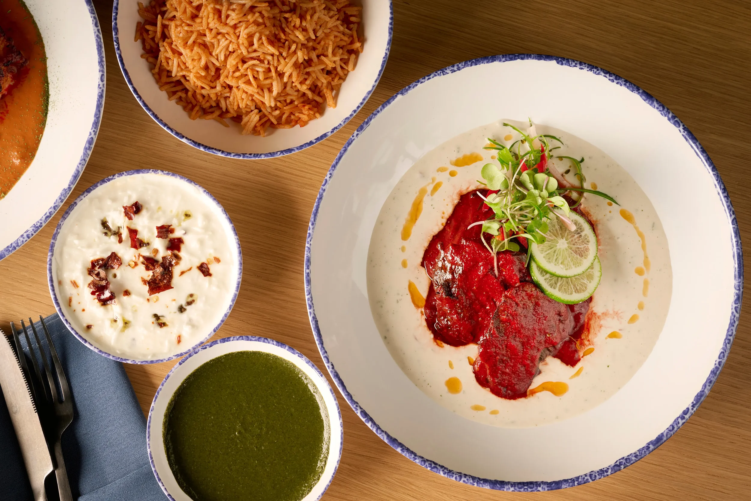 Overhead view of a white plate with Spicy Rajasthani Red Chili Lamb Masala and sides of rice, cucumber raita, and green herb sauce from the Samudra in Farewinds Food Hall.