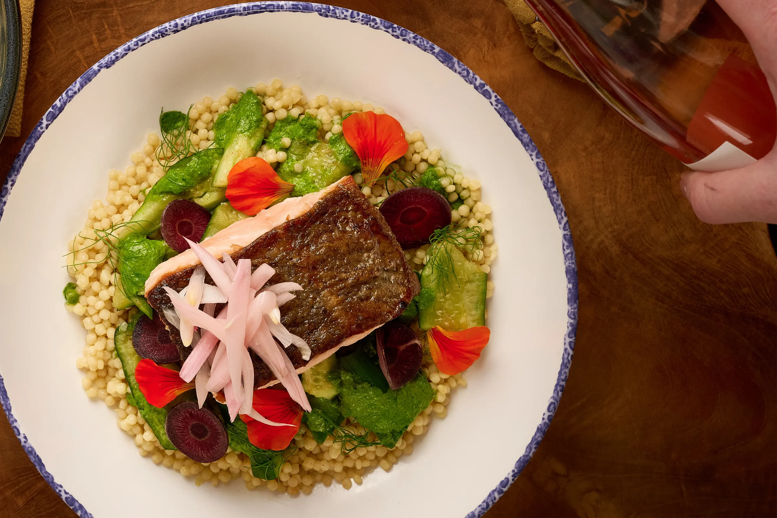 Overhead view of a white plate with herb crusted salmon over hand-rolled semolina couscous, green chermoula, anchovy, and pickled fennel from the Mezza slip in Farewinds Food Hall.