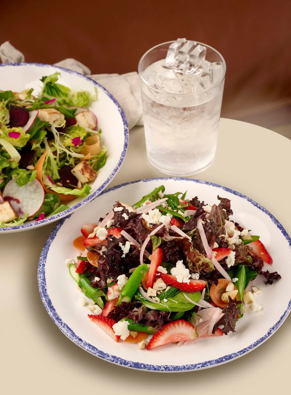 Two white plates, one with a strawberry and asparagus salad and the other with a beet and halloumi salad, next to a glass of ice water from the Mezza slip in Farewinds Market Hall.