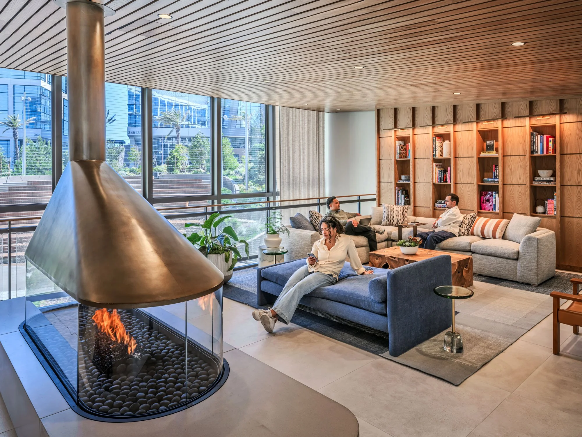 Customers relaxing on couches in front of modern bookshelves in the reservable lounge, with a glass-walled fireplace and hood and window walls looking out onto a landscaped outdoor area.