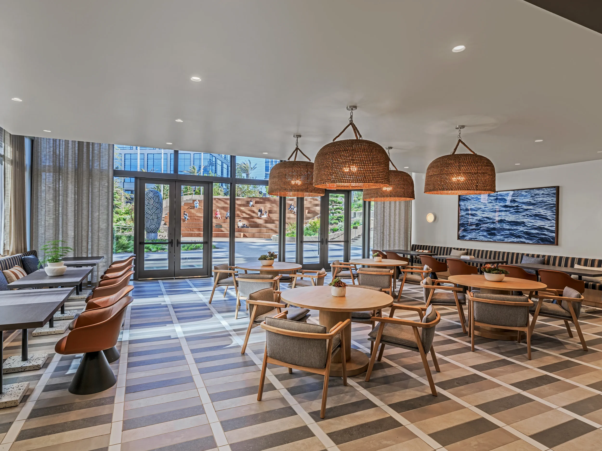 A bright, reservable dining room at Farewinds with round tables with modern chairs in the center, padded benches with square tables along the walls, wicker pendant lights, and window-wall opening onto a landscaped outdoor area.