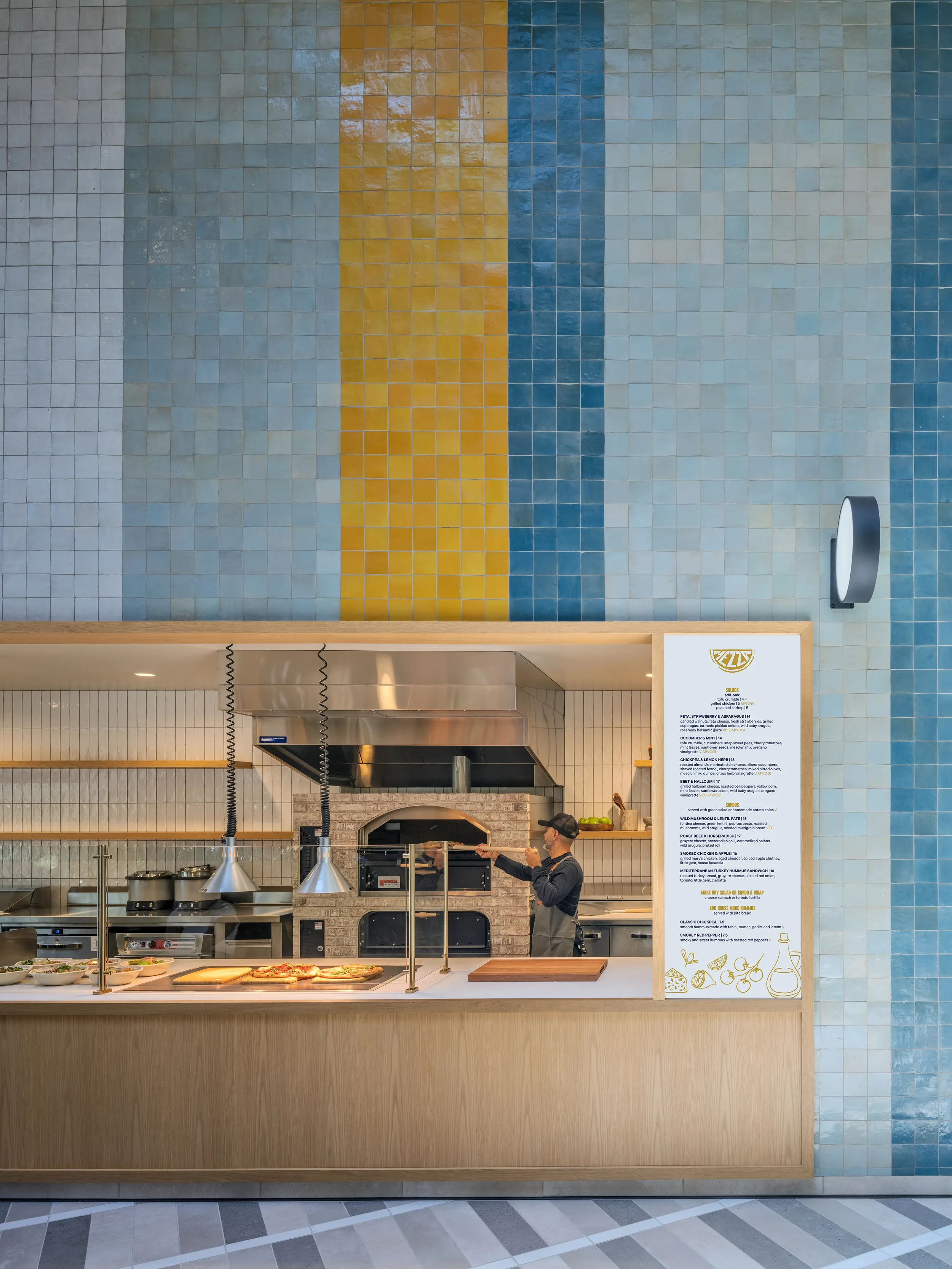 The Mezza slip at Farewinds Food Hall where an employee is removing a grilled pita from a large brick oven, with food displayed at the counter for pick-up and a large menu displayed to the right.