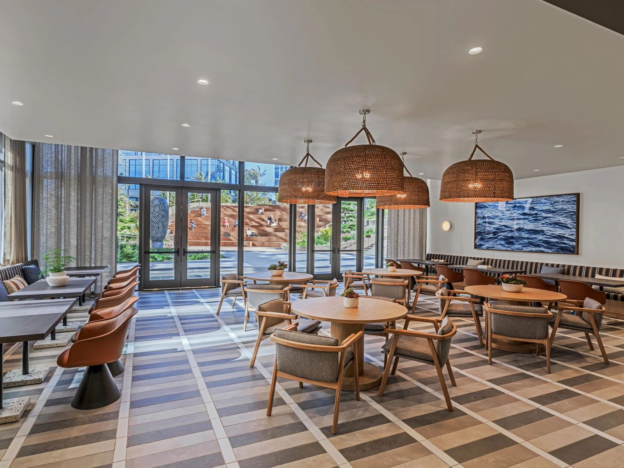 A bright, reservable dining room at Farewinds with round tables with modern chairs in the center, padded benches with square tables along the walls, wicker pendant lights, and window-wall opening onto a landscaped outdoor area.