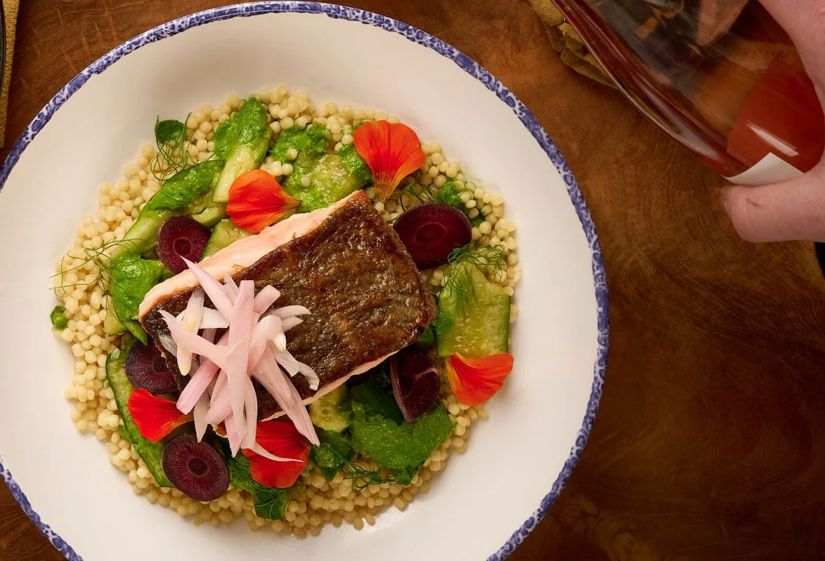 Overhead view of a white plate with herb crusted salmon over hand-rolled semolina couscous, green chermoula, anchovy, and pickled fennel from the Mezza slip in Farewinds Market Hall.
