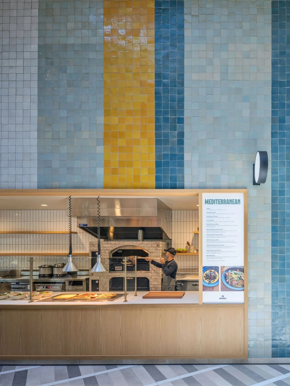 The Mezza slip at Farewinds Market Hall where an employee is removing a grilled pita from a large brick oven, with food displayed at the counter for pick-up and a large menu displayed to the right.