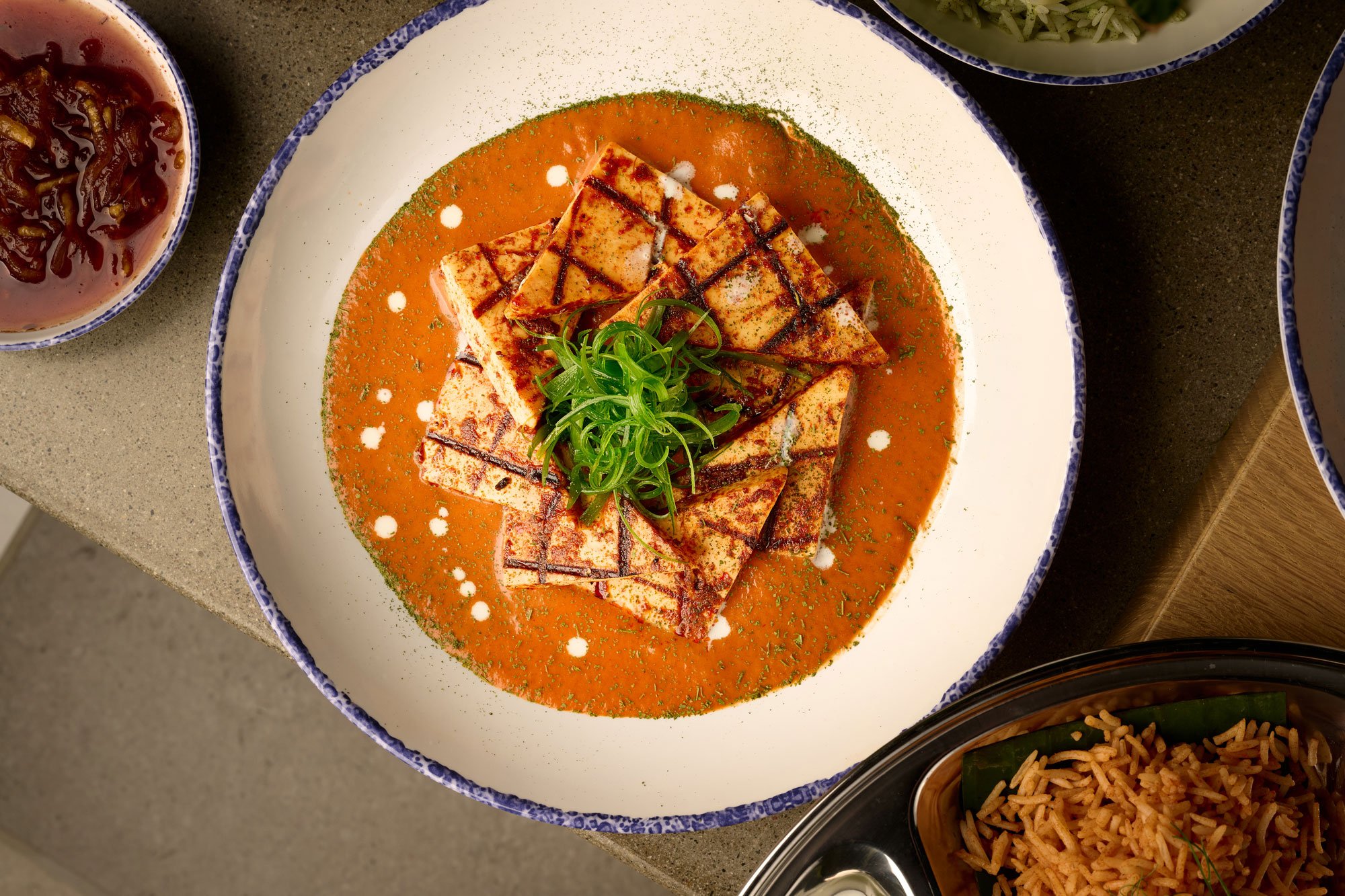 Overhead view of a white bowl with paneer tikka masala, served with triangles of grilled indian paneer cheese over smoked tomato and cashew yogurt cream sauce from the Indian slip in Farewinds Market Hall.