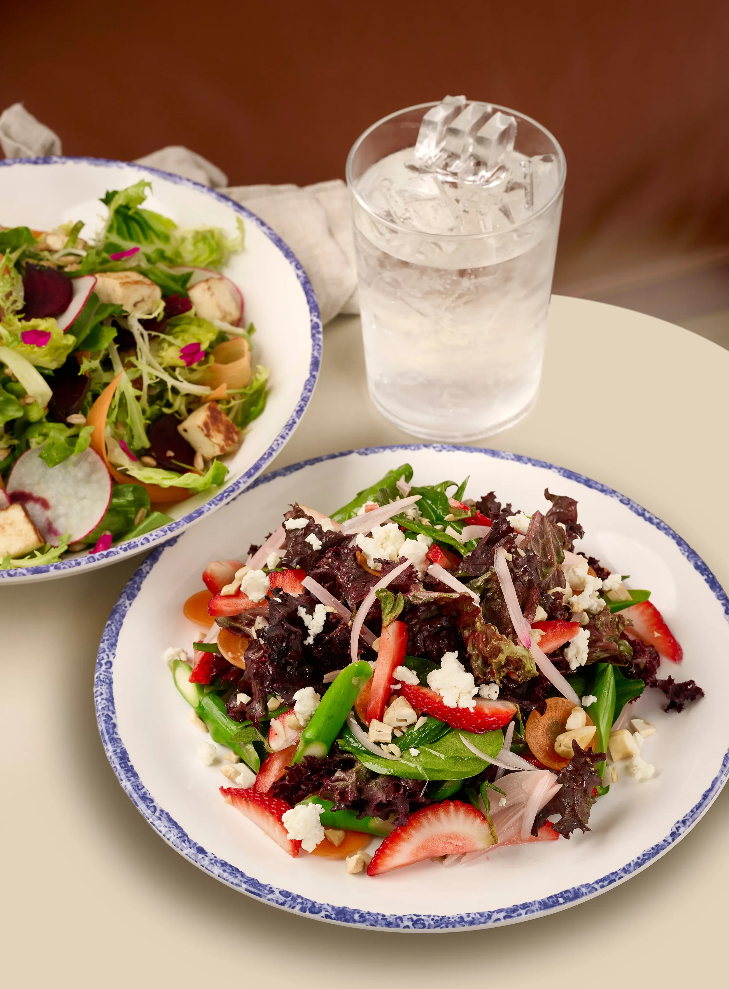 Two white plates, one with a strawberry and asparagus salad and the other with a beet and halloumi salad, next to a glass of ice water from the Mezza slip in Farewinds Food Hall.
