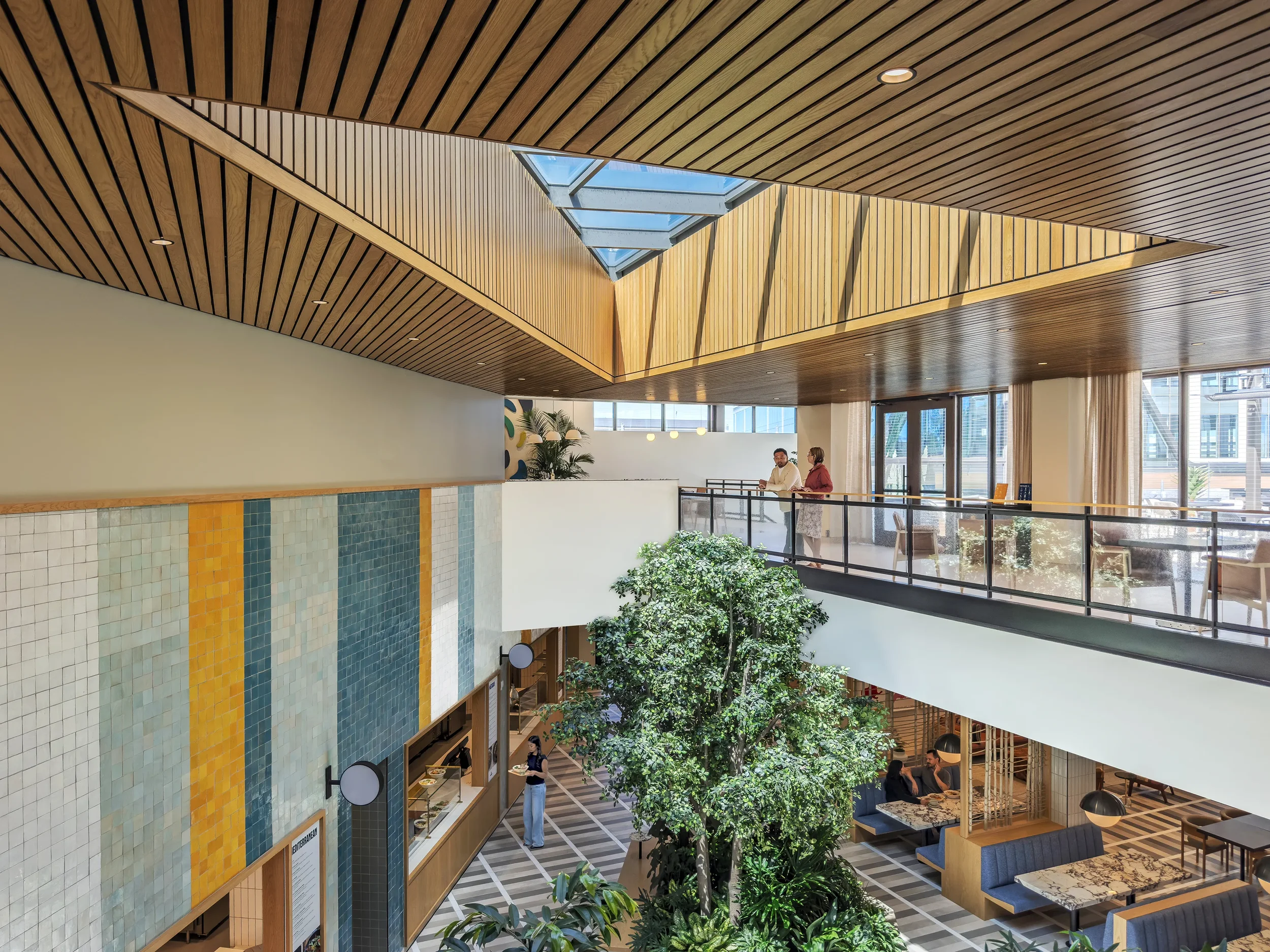 Two people stand on a second floor interior balcony at Farewinds overlooking a two-story atrium with large plants and a triangle sunroof, with dining booths visible on the first floor.