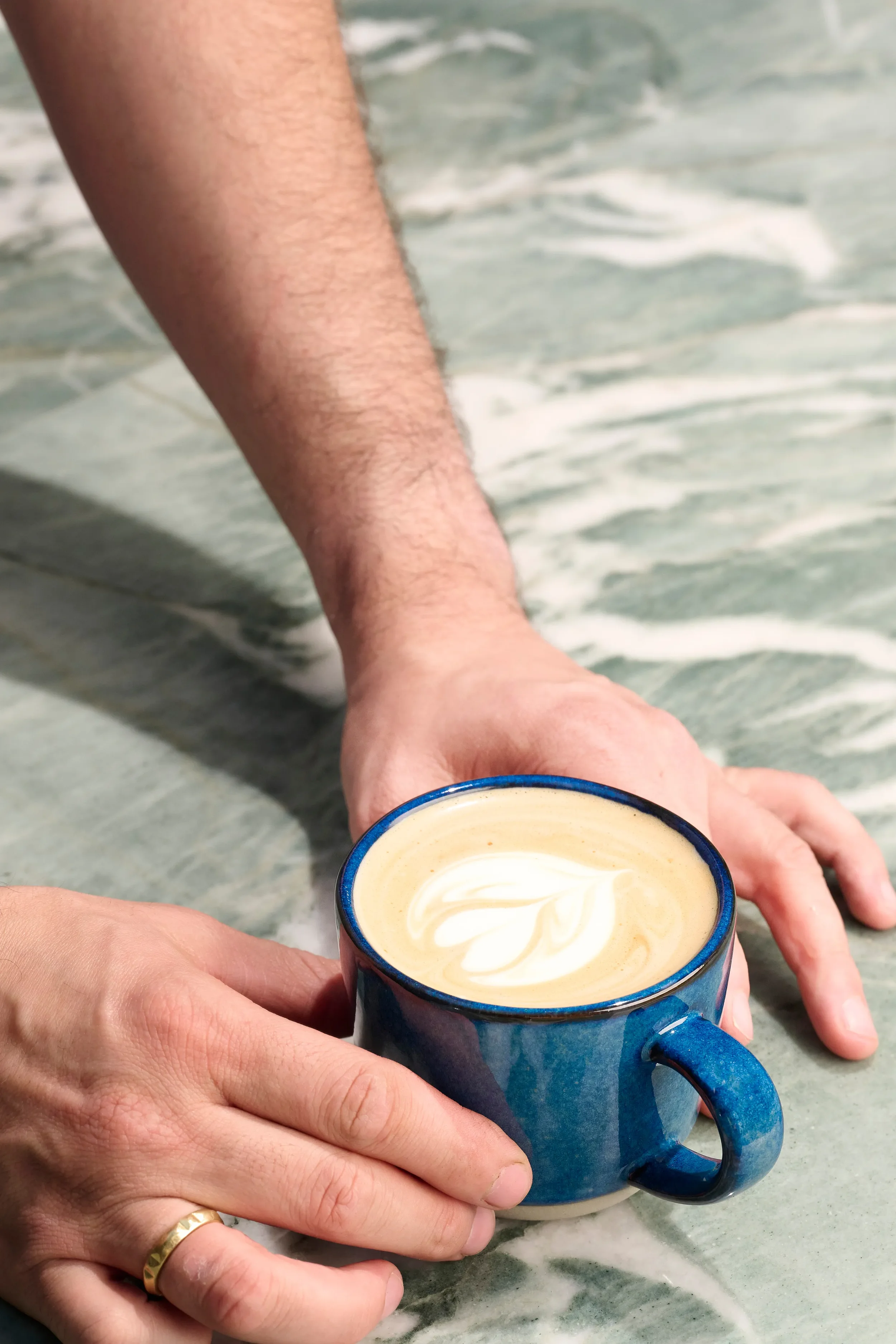 Close-up of two hands setting a freshly made latte in a blue ceramic cup on the counter at Farewinds Market.