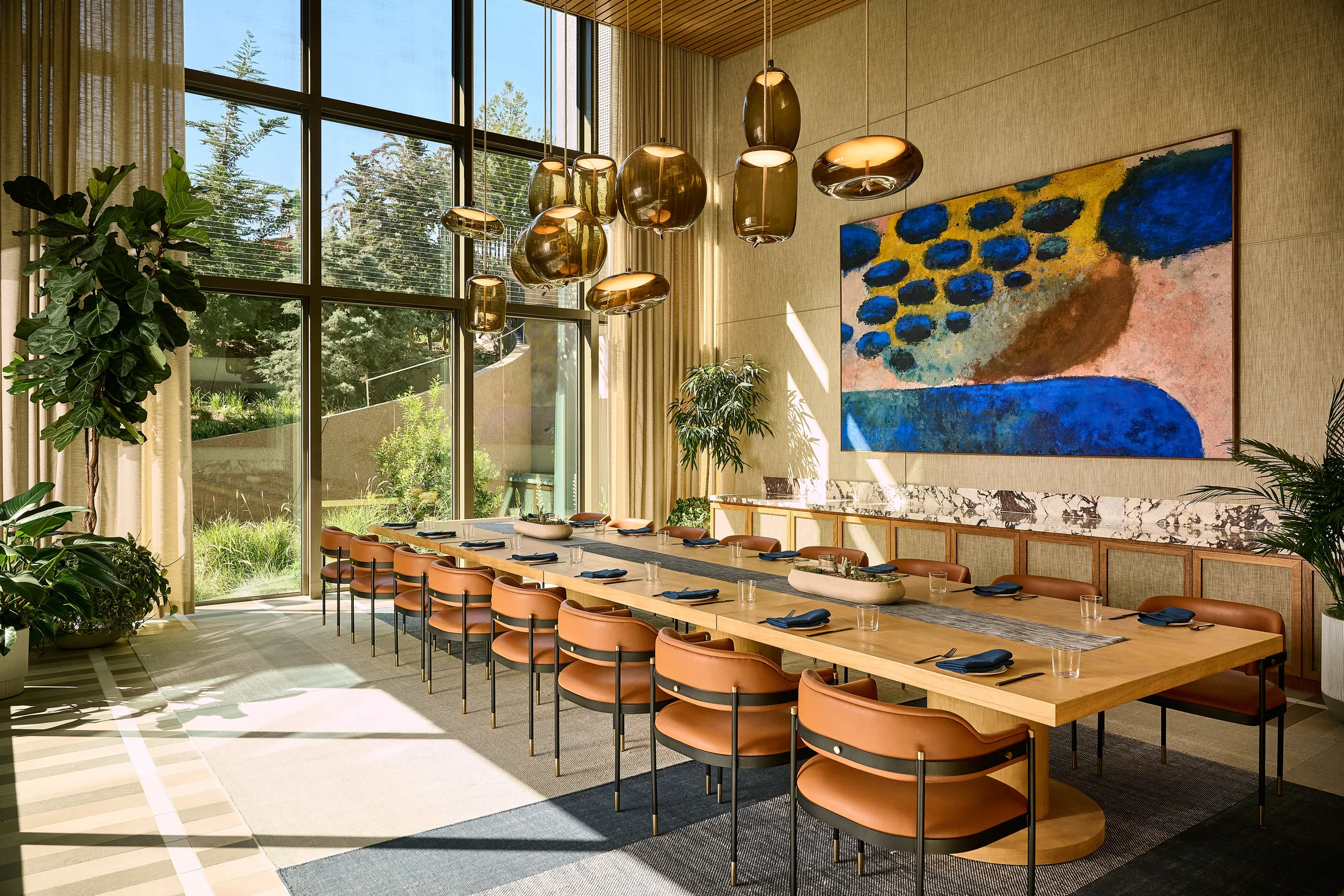 Reservable private dining room at Farewinds with AV system, long wooden table, brown chairs, large windows, plants, and colorful abstract wall art.