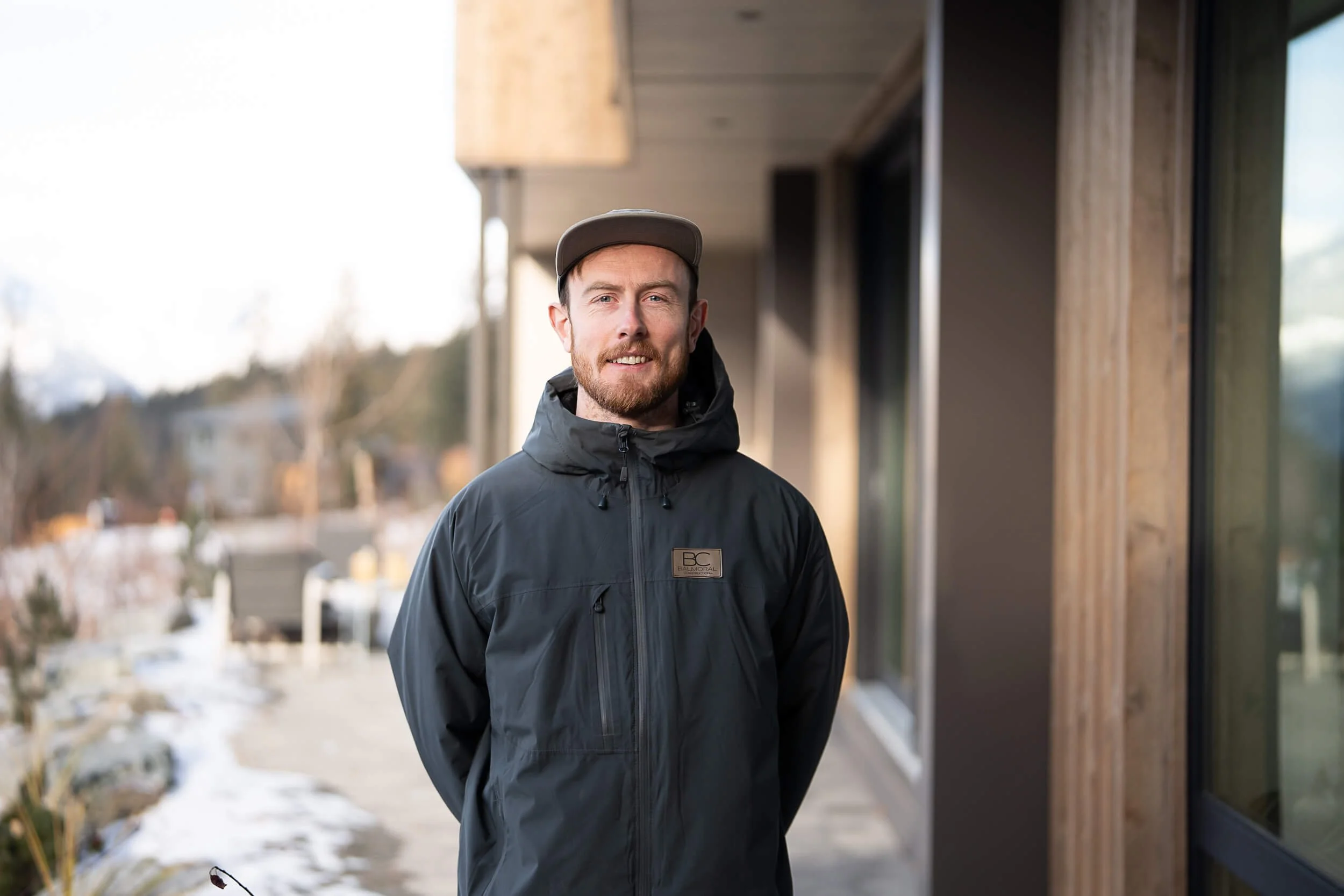Man in a black jacket and hat standing outdoors near a building.