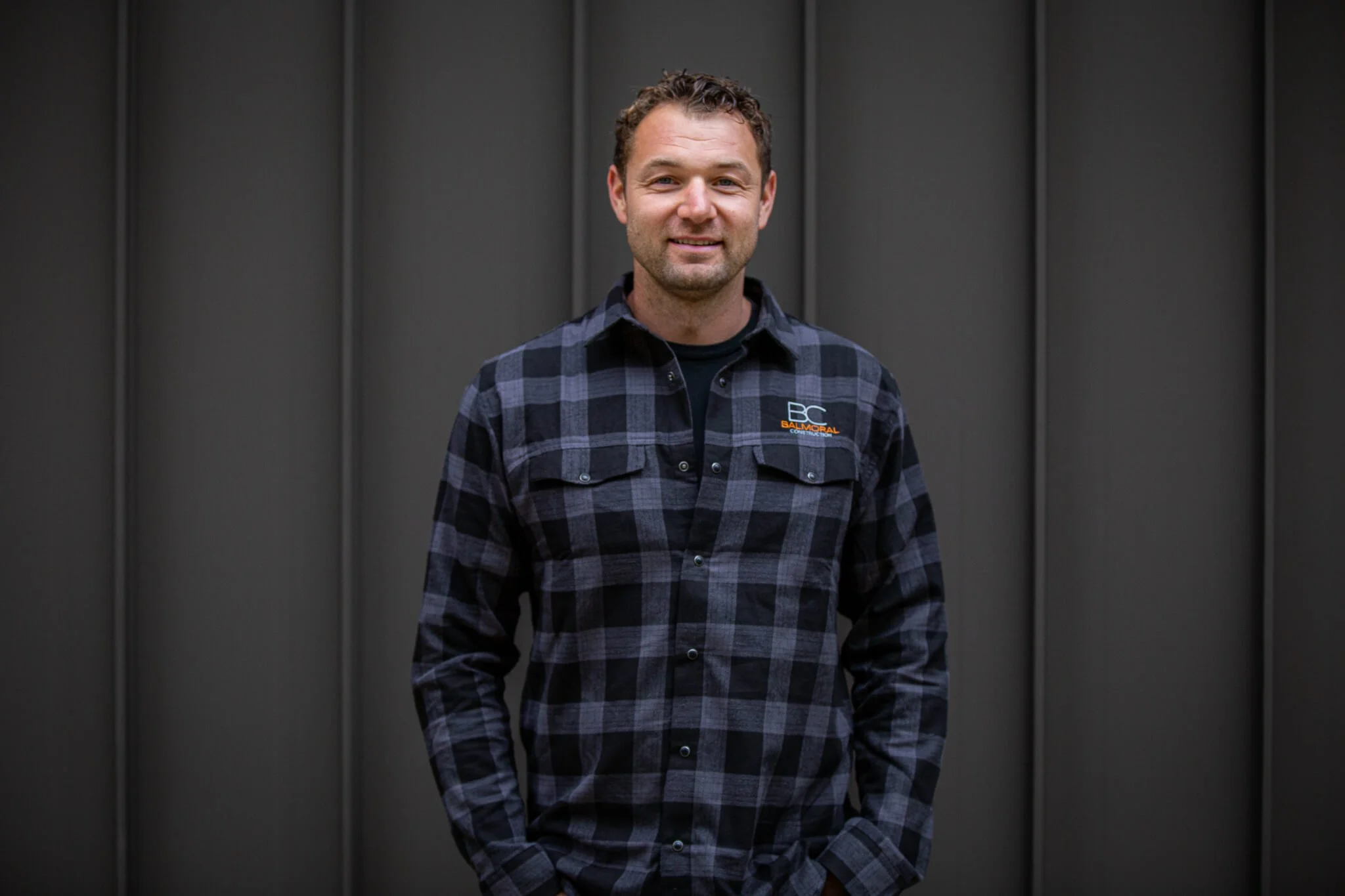 Person wearing a plaid shirt with "BC Balmoral" logo, standing against a dark, textured background.