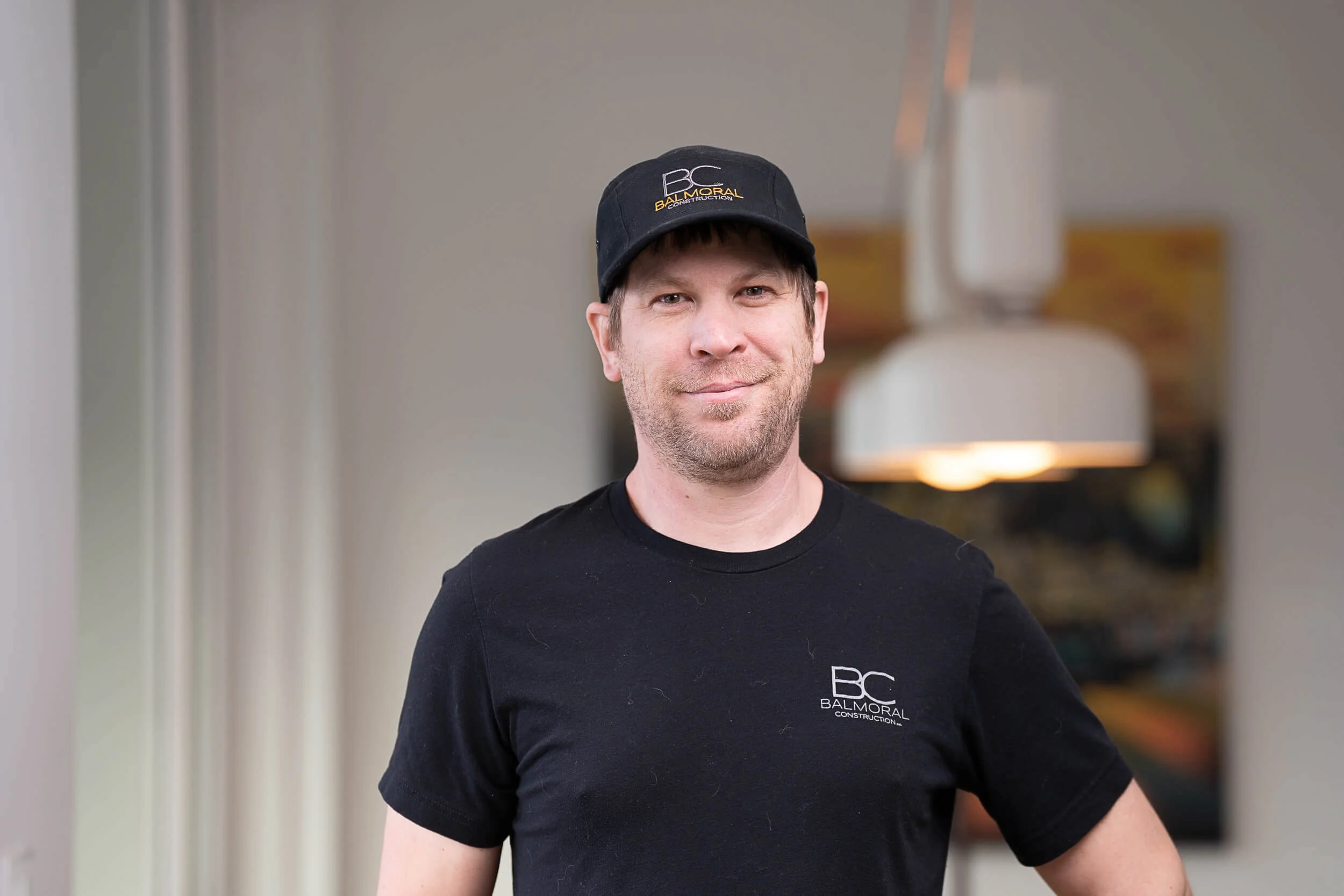 Man wearing a black cap and black shirt with "Balmoral Construction" logo, standing indoors with a light fixture and blurred background.