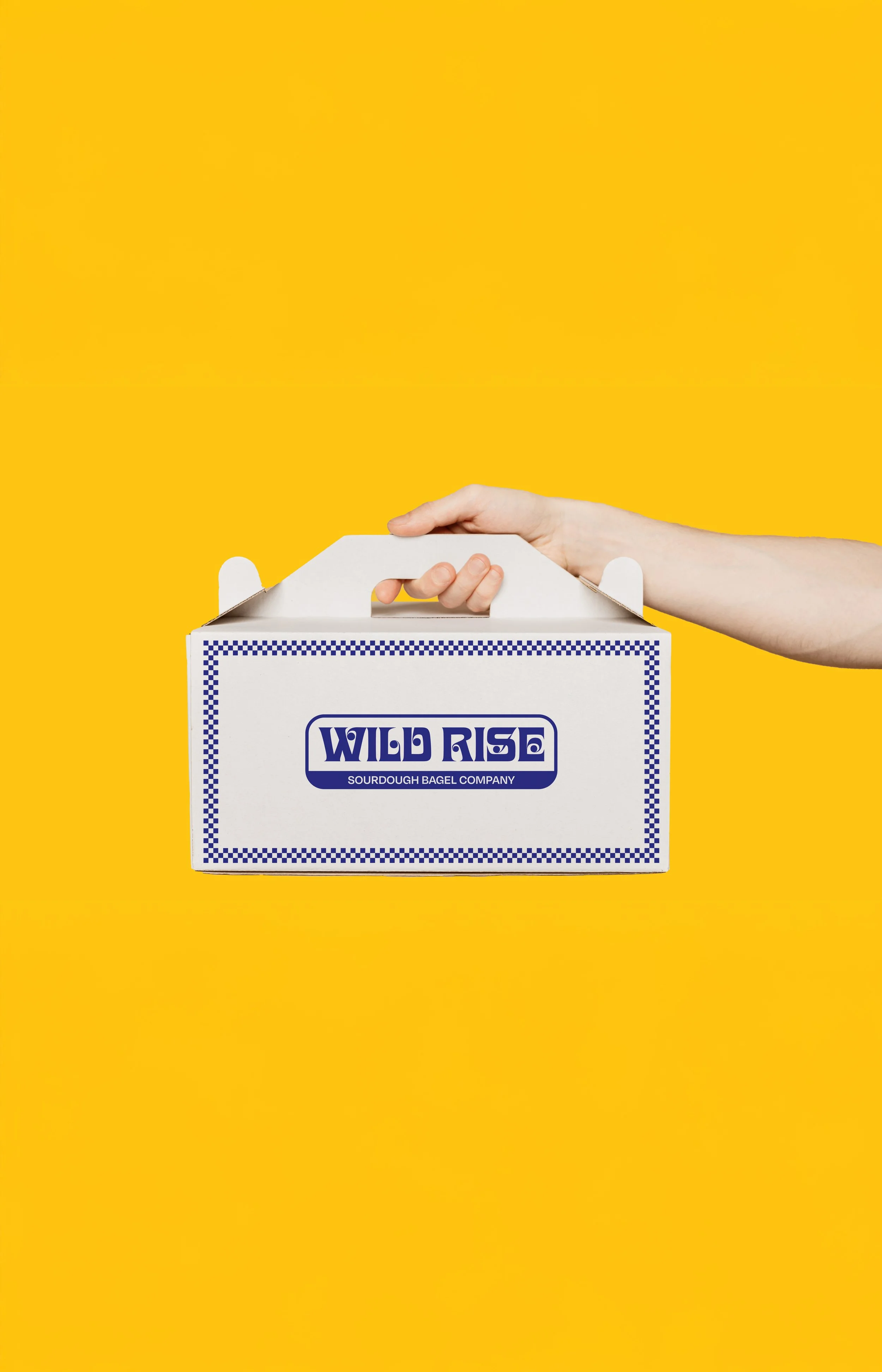 A hand holding a white box with blue text that reads 'Wild Rise Sourdough Bagel Company' against a yellow background.