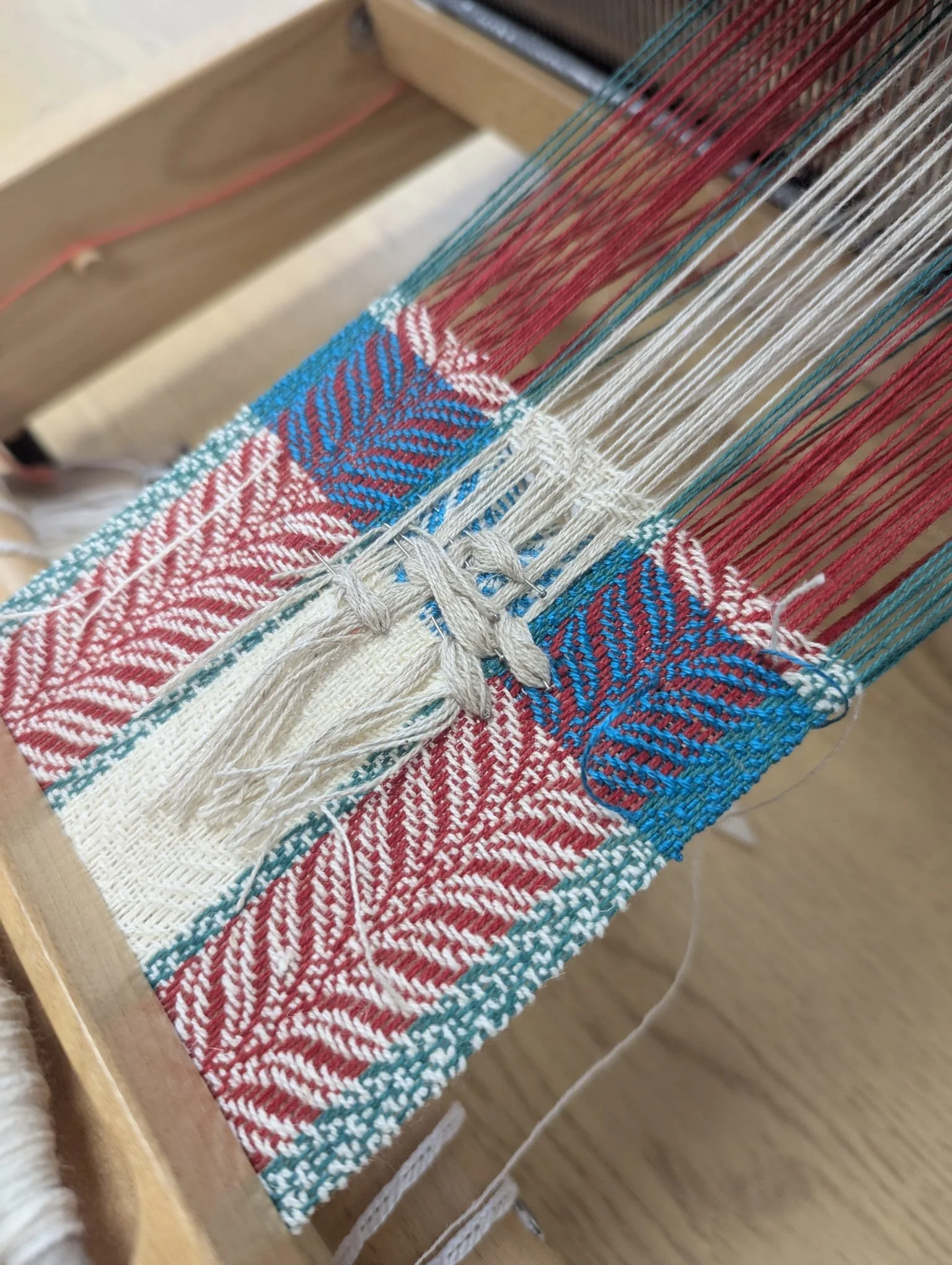 Close-up of a colorful woven textile on a loom, with red, blue, and white threads in a detailed leaf and branch pattern.