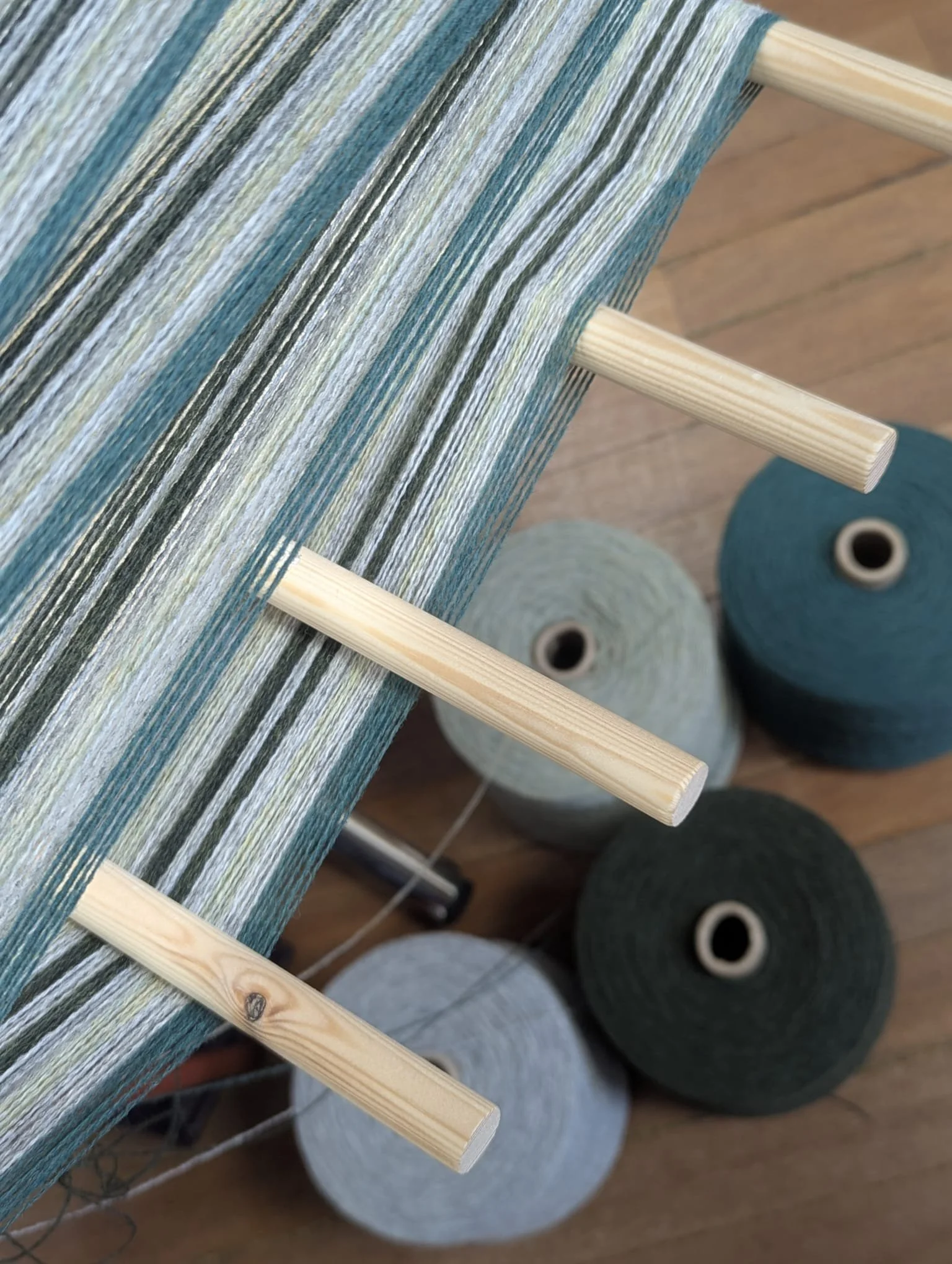 Close-up of a loom with multi-colored yarn strings and several large spools of yarn underneath on a wooden floor.