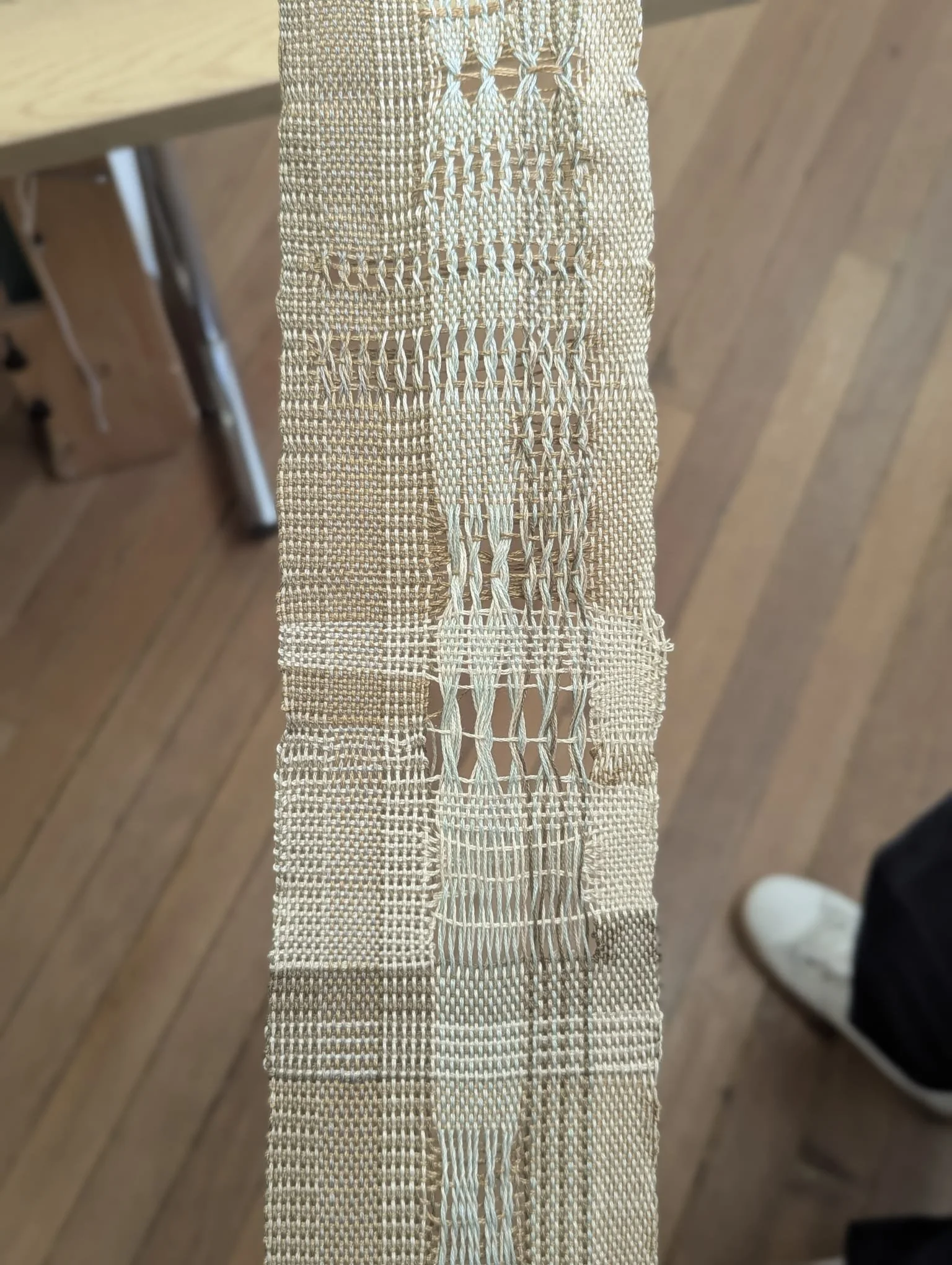 Close-up of a woven, beige lace fabric with intricate open-weave patterns, placed on a wooden surface with a person’s leg and shoe visible in the background.