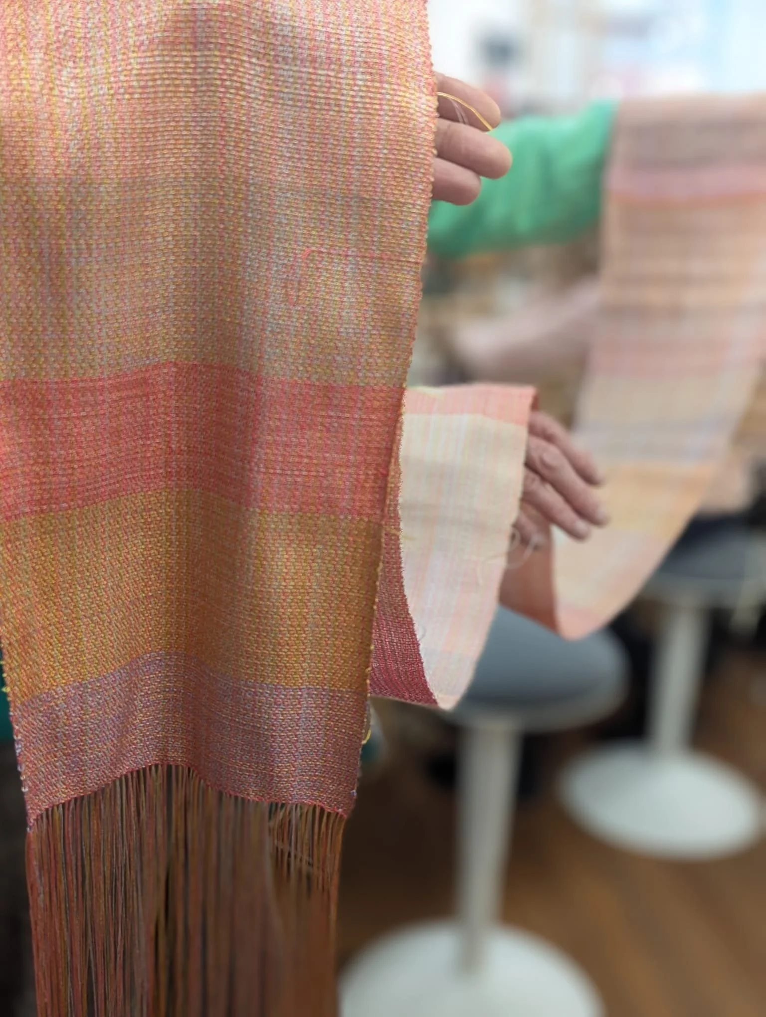 Close-up of woven fabric swatches in pastel colors, held by hands with rings, with a blurred background of more fabric samples on display.