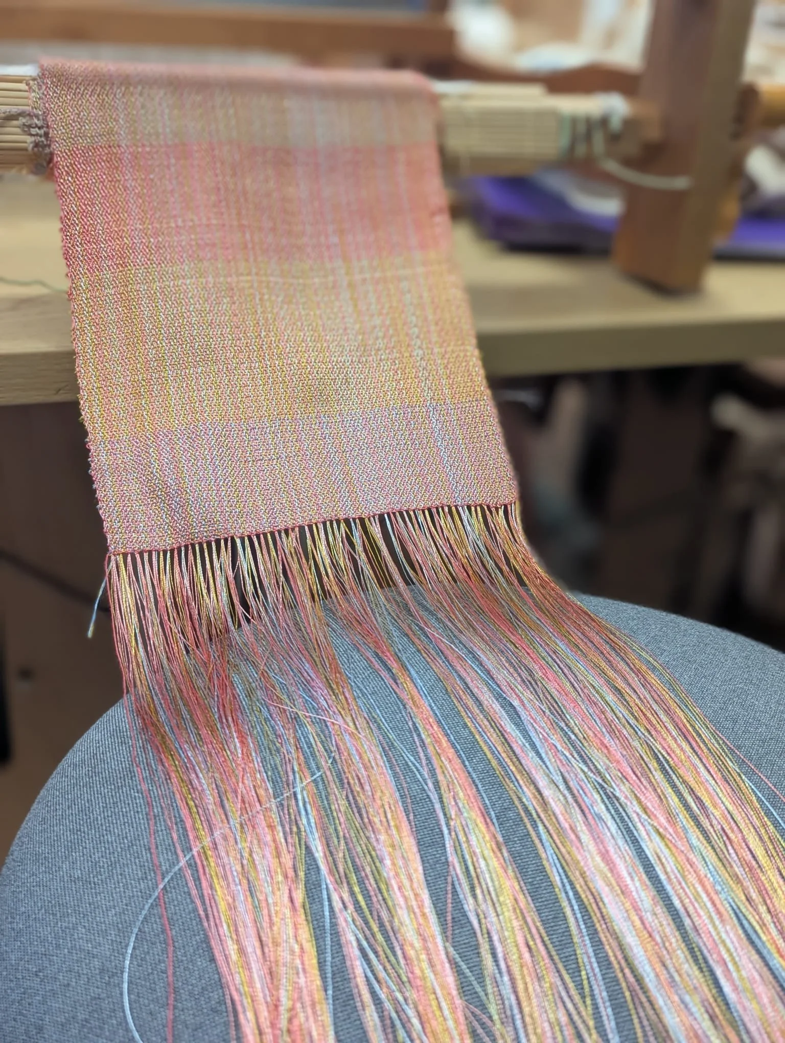 Close-up of a weaving loom with a partially woven textile in pastel pink and yellow shades, with loose threads hanging from the loom.