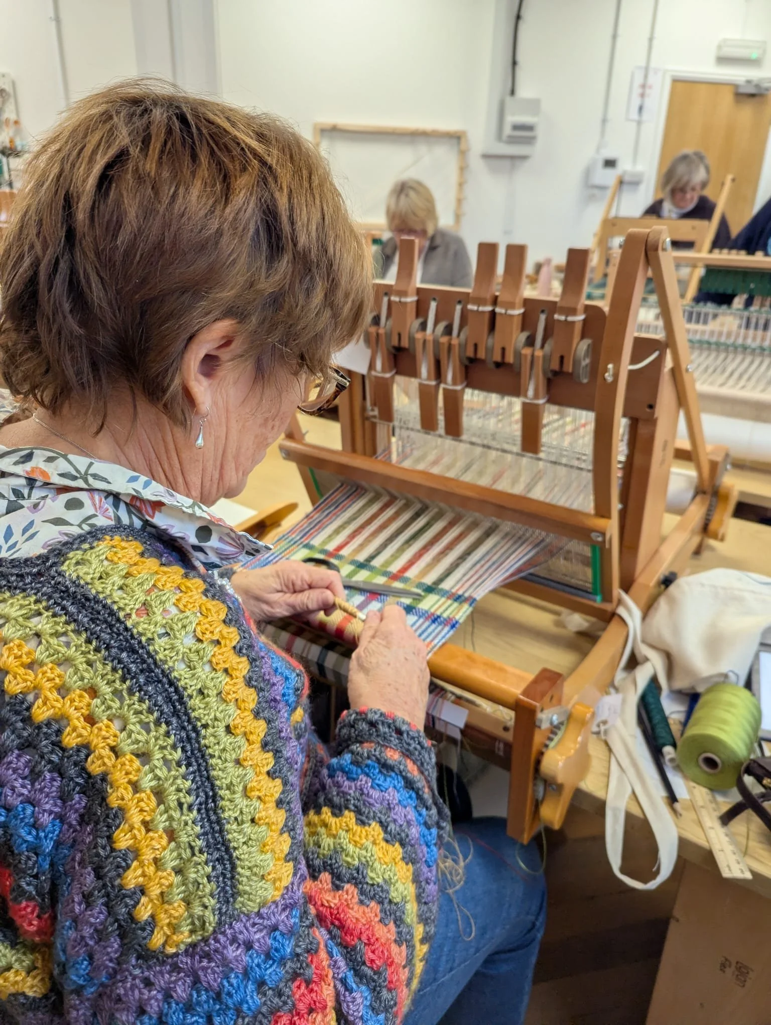 An elderly woman in a multicolored crocheted sweater weaving on a tapestry loom in a craft workshop.