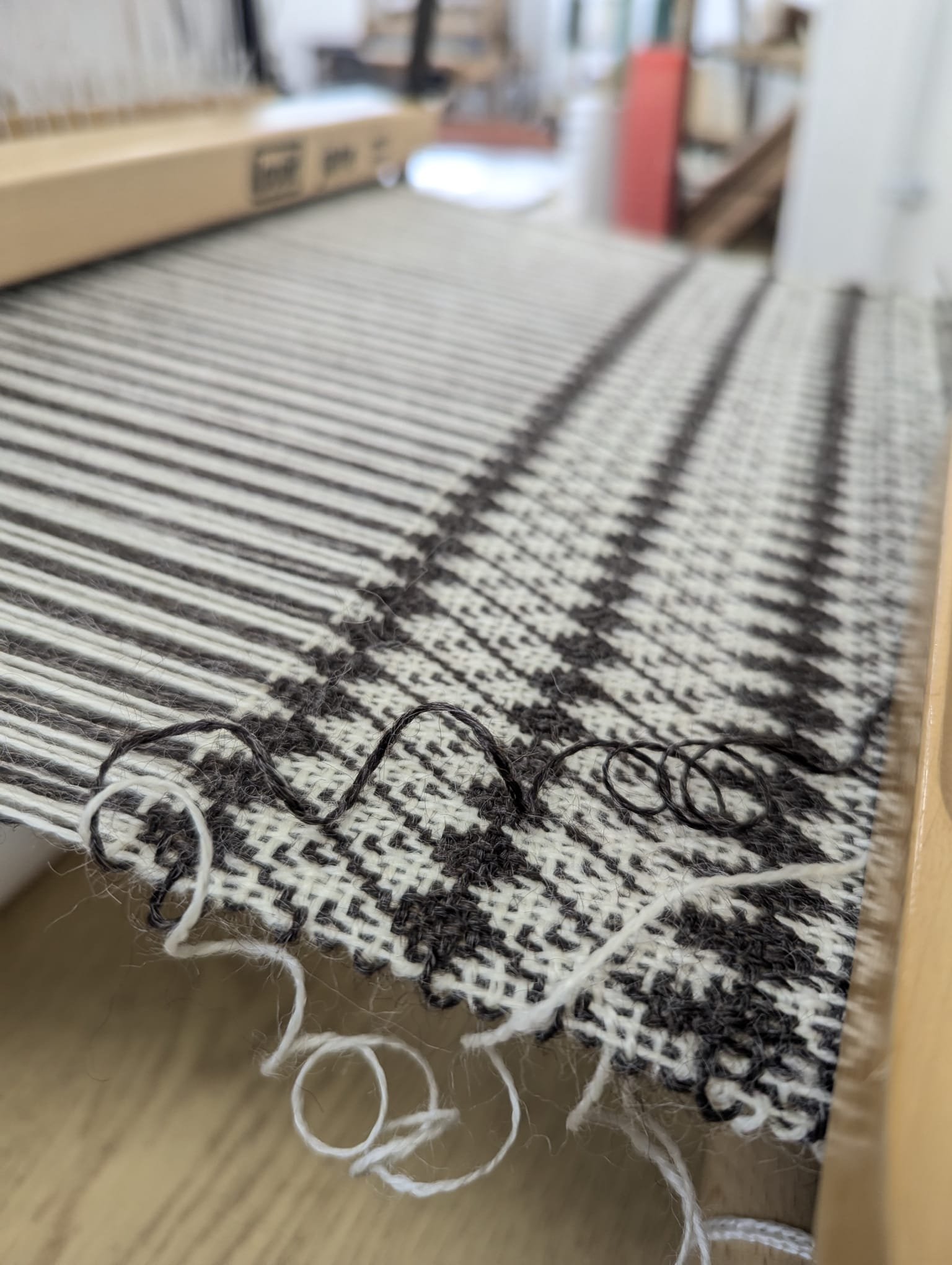 Close-up of a weaving loom with black and white yarn, partially woven fabric, and loose yarn loops hanging from the edge.