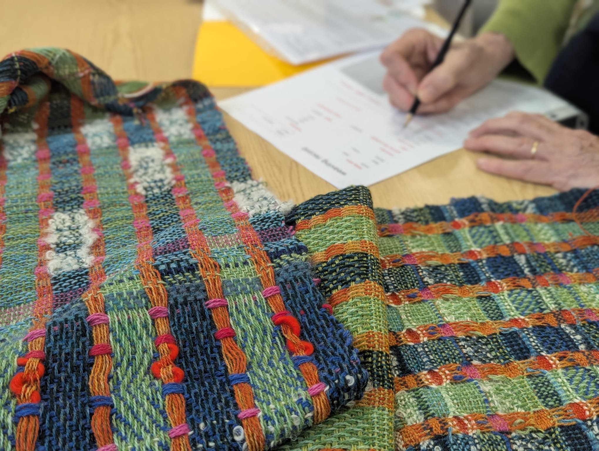 Colorful woven fabric with vibrant patterns on a wooden table, alongside a person in a green jacket writing on a piece of paper.