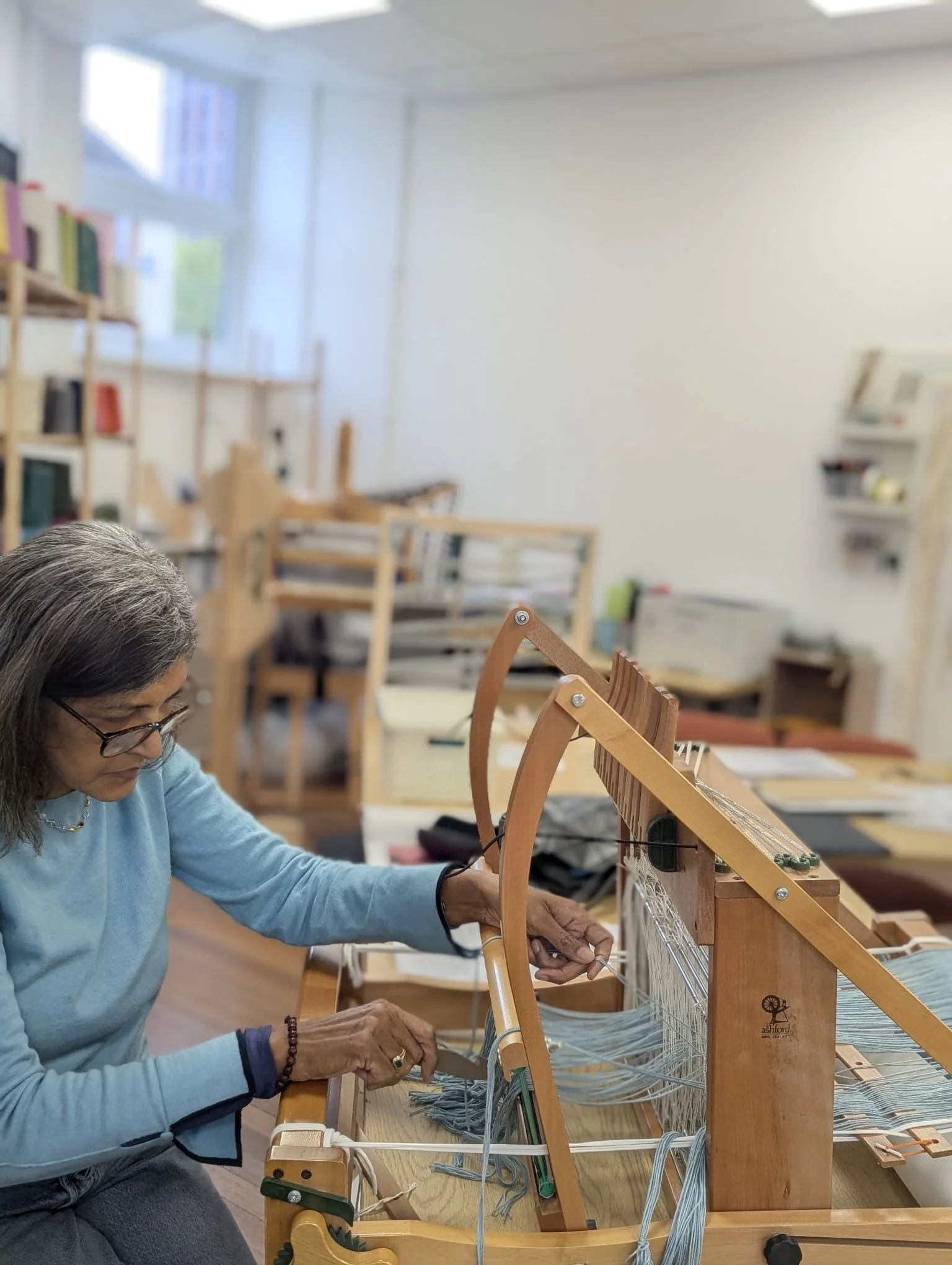 An elderly woman wearing glasses and a light blue sweater working on a wooden loom in a well-lit room with white walls and shelves.
