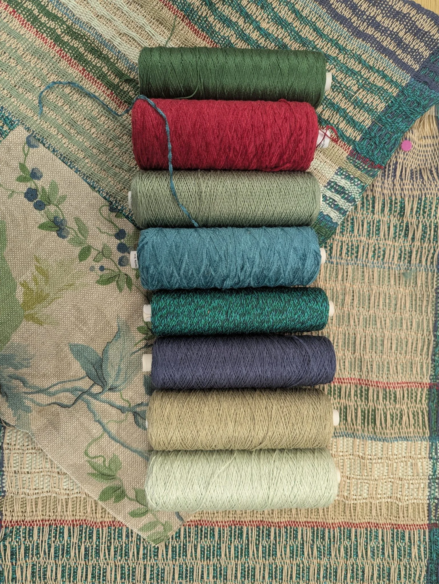 Ten spools of colored thread arranged vertically on a multicolored woven fabric background.