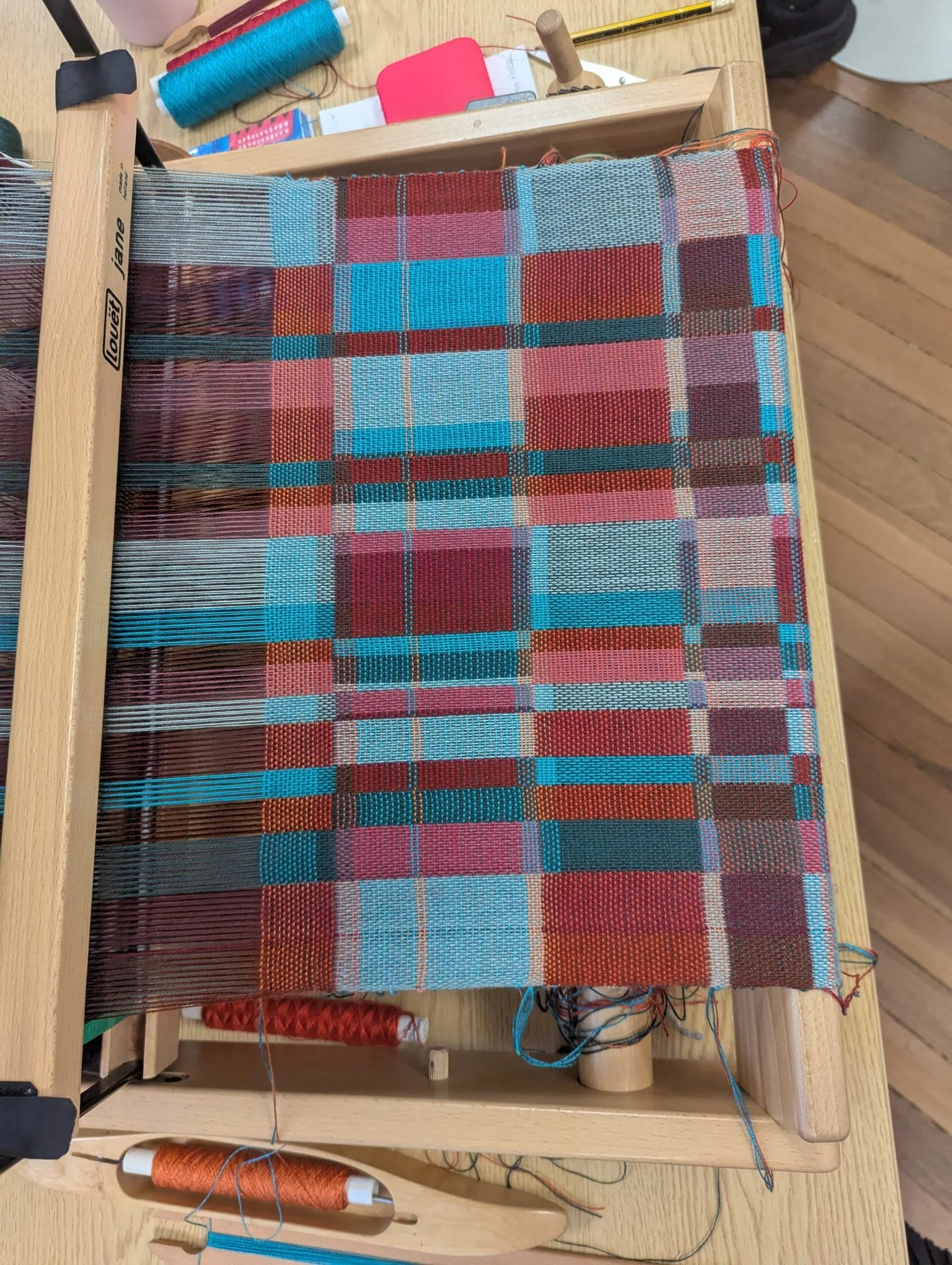 A weaving loom with a partially woven colorful plaid fabric in red, blue, and pink tones, surrounded by spools of thread, a shuttle, and other weaving tools on a wooden table.