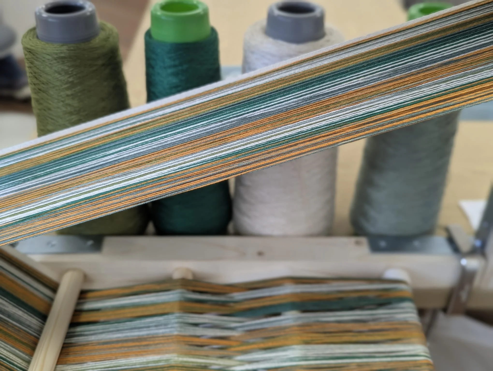 Multiple spools of thread in various shades of green, blue, gray, and beige, with some threads wound onto stacked paper or cardboard, on a weaving or sewing machine.