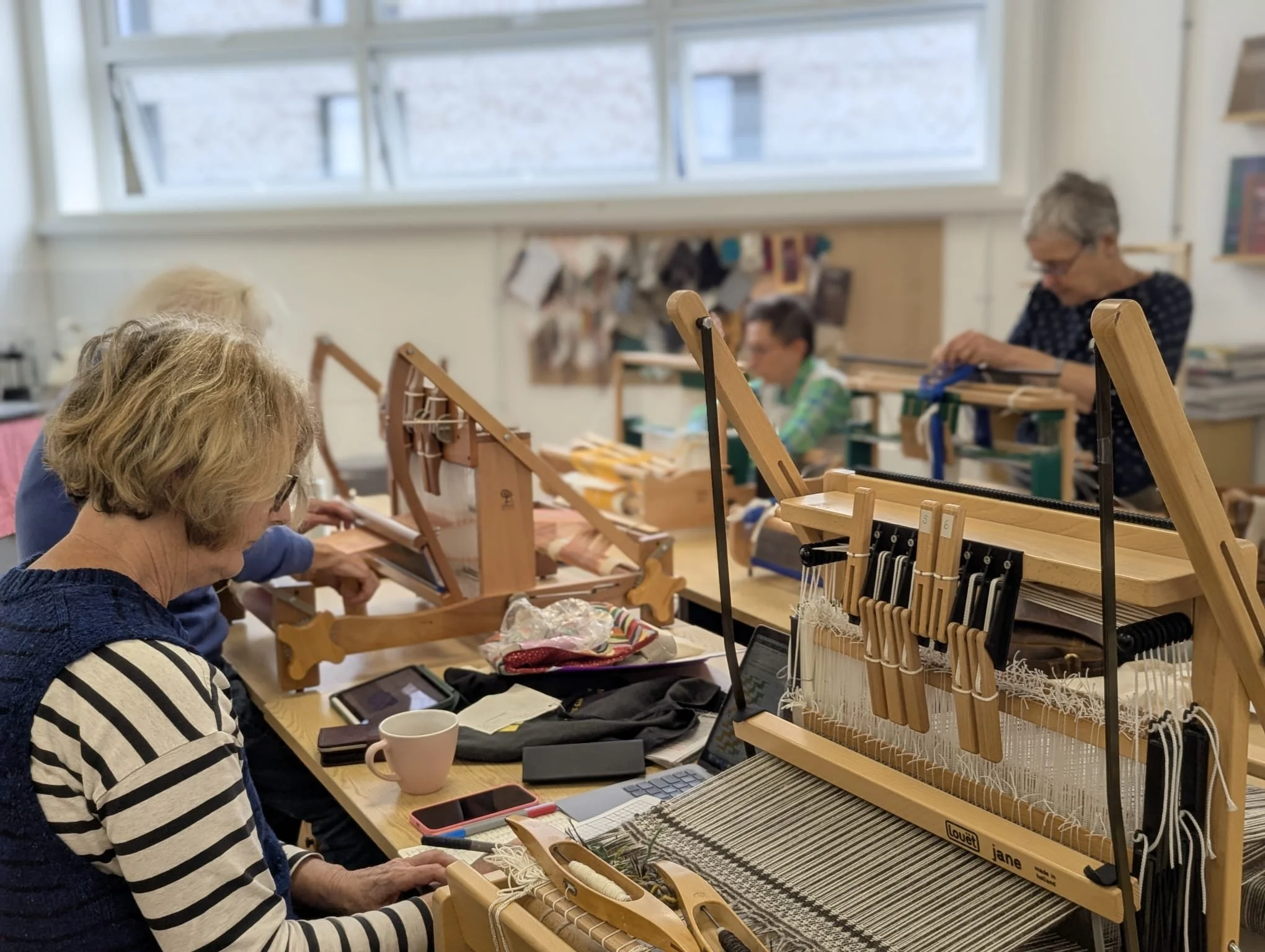 A group of women engaging in loom weaving in a bright room with large windows, wooden weaving looms, and various weaving tools on a table.