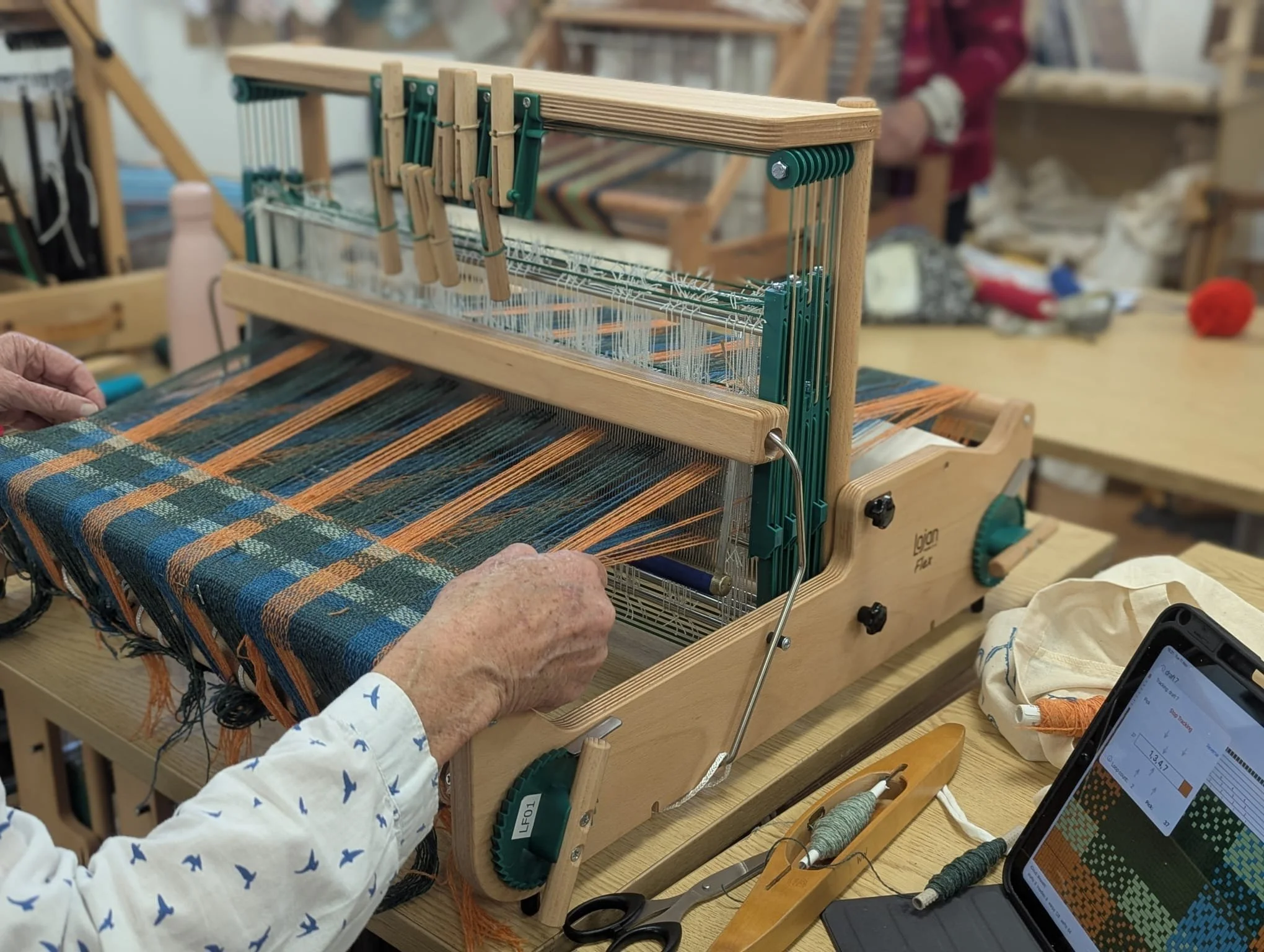 An elderly person weaves a plaid fabric on a wooden loom in a craft room. Various weaving tools and a tablet are on the table.
