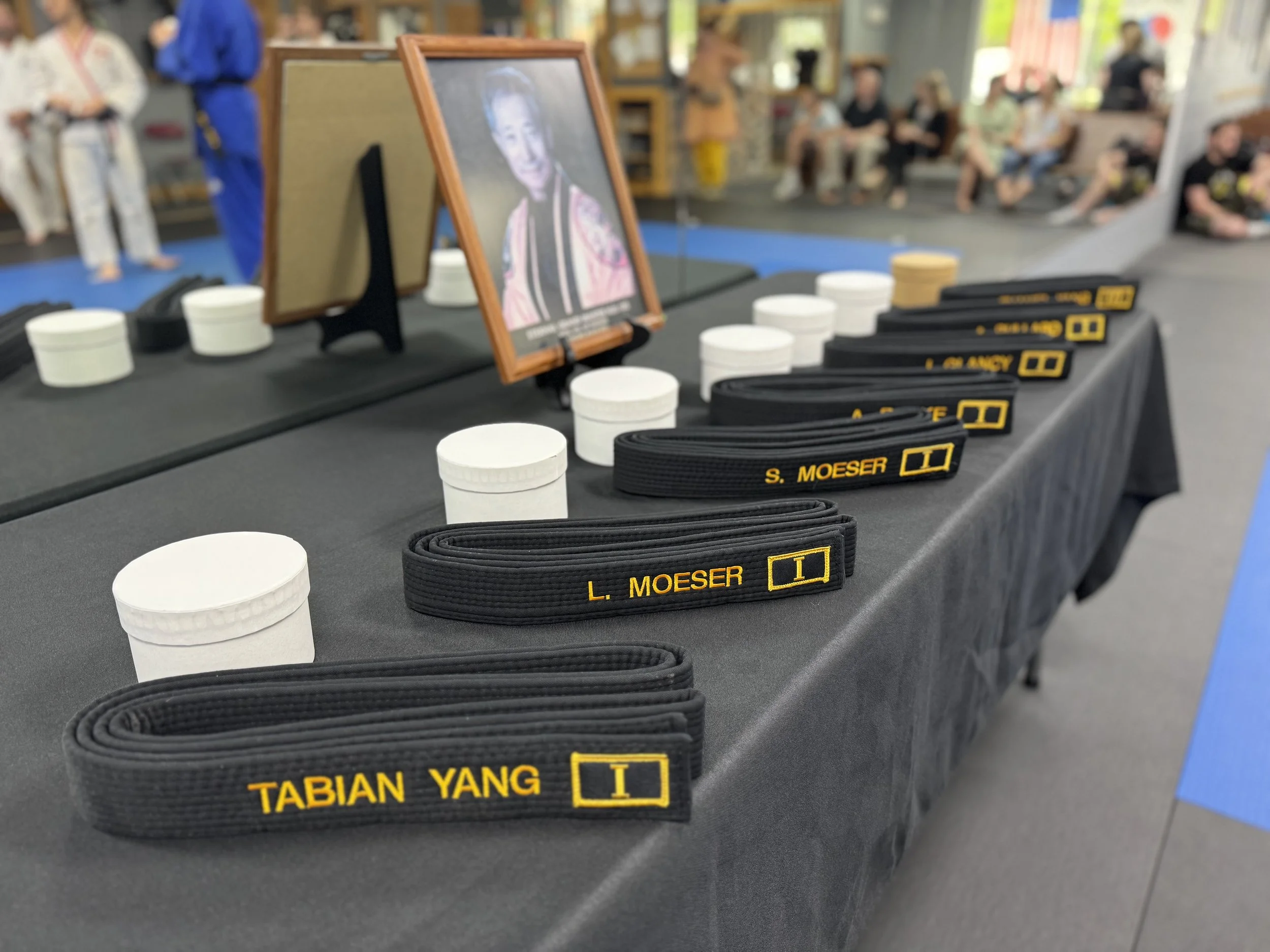 Table with martial arts belts, white cups, and a framed photo. People in martial arts uniforms and casual clothing are seated in the background.