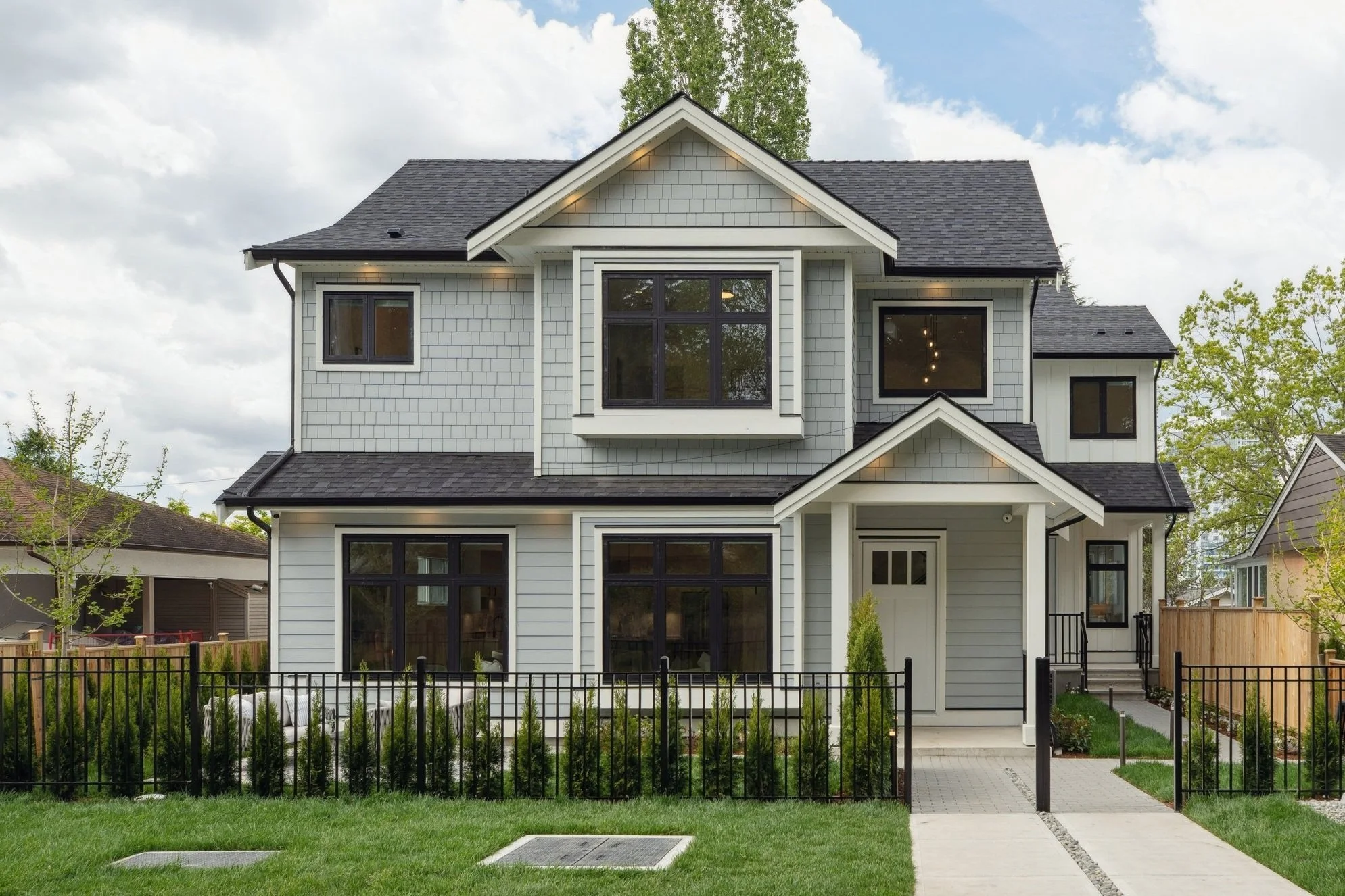 A modern, multi-story house with gray siding, black window frames, and a gabled roof, surrounded by a black metal fence and a lush green lawn.