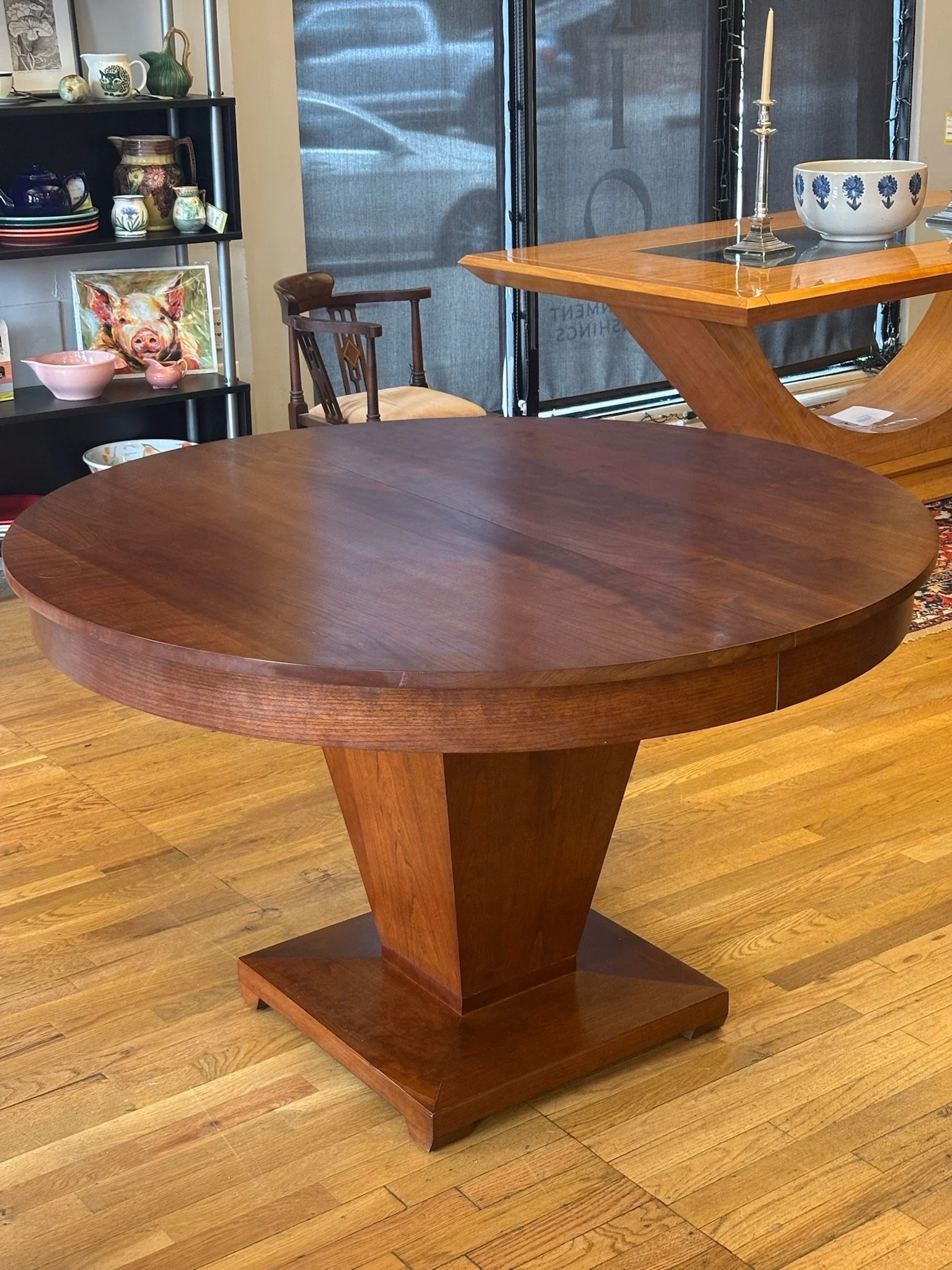 A wooden dining table with a thick round top and a pedestal base in a furniture store. There is a chair behind the table, a black shelf with pottery and art in the background, and a decorative bowl with a candle and a ceramic bowl on a glass table in