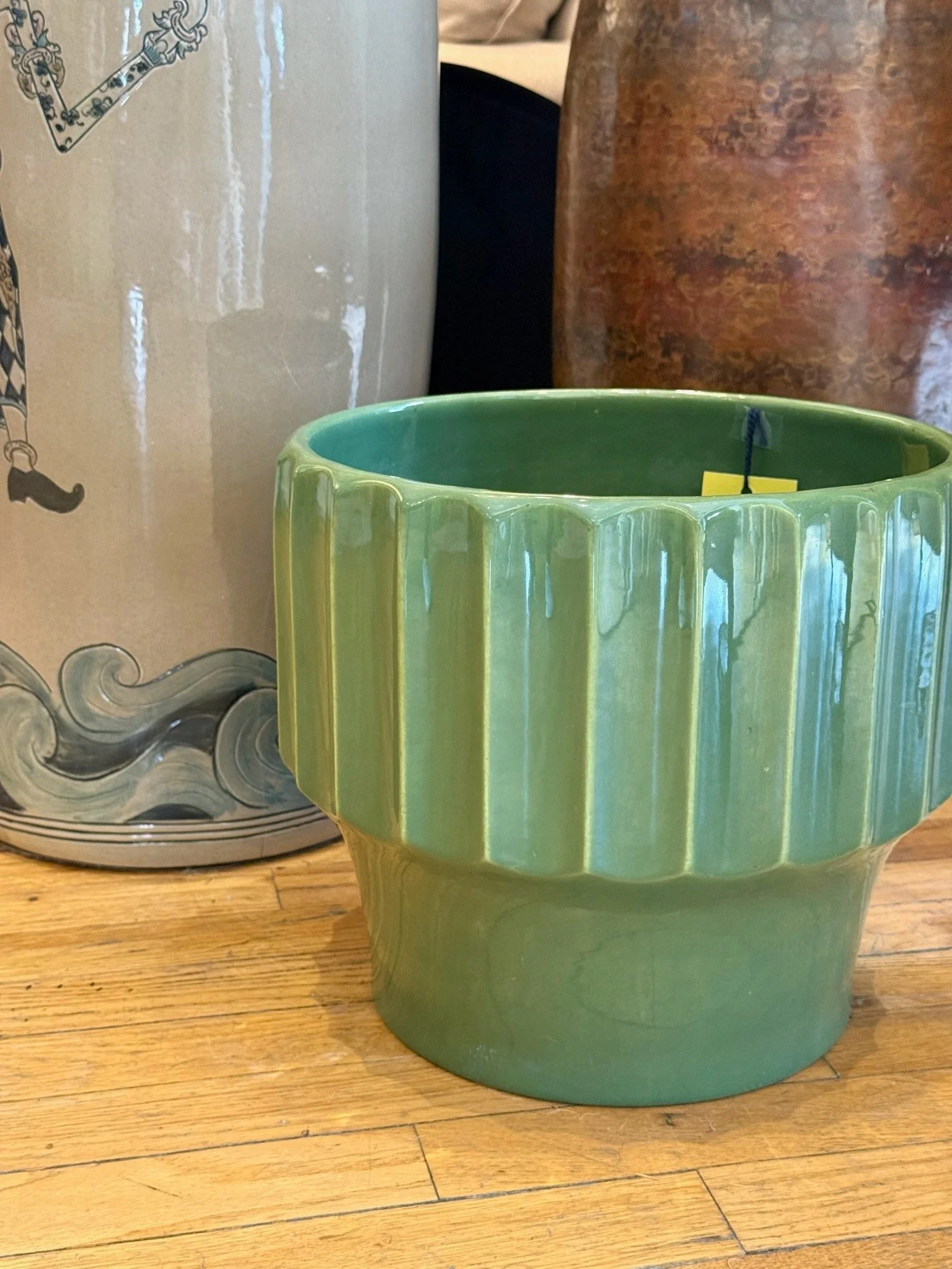 A green ceramic planter with a ridged design on the outside, placed on a wooden floor next to a grey cylindrical object with a decorative pattern.