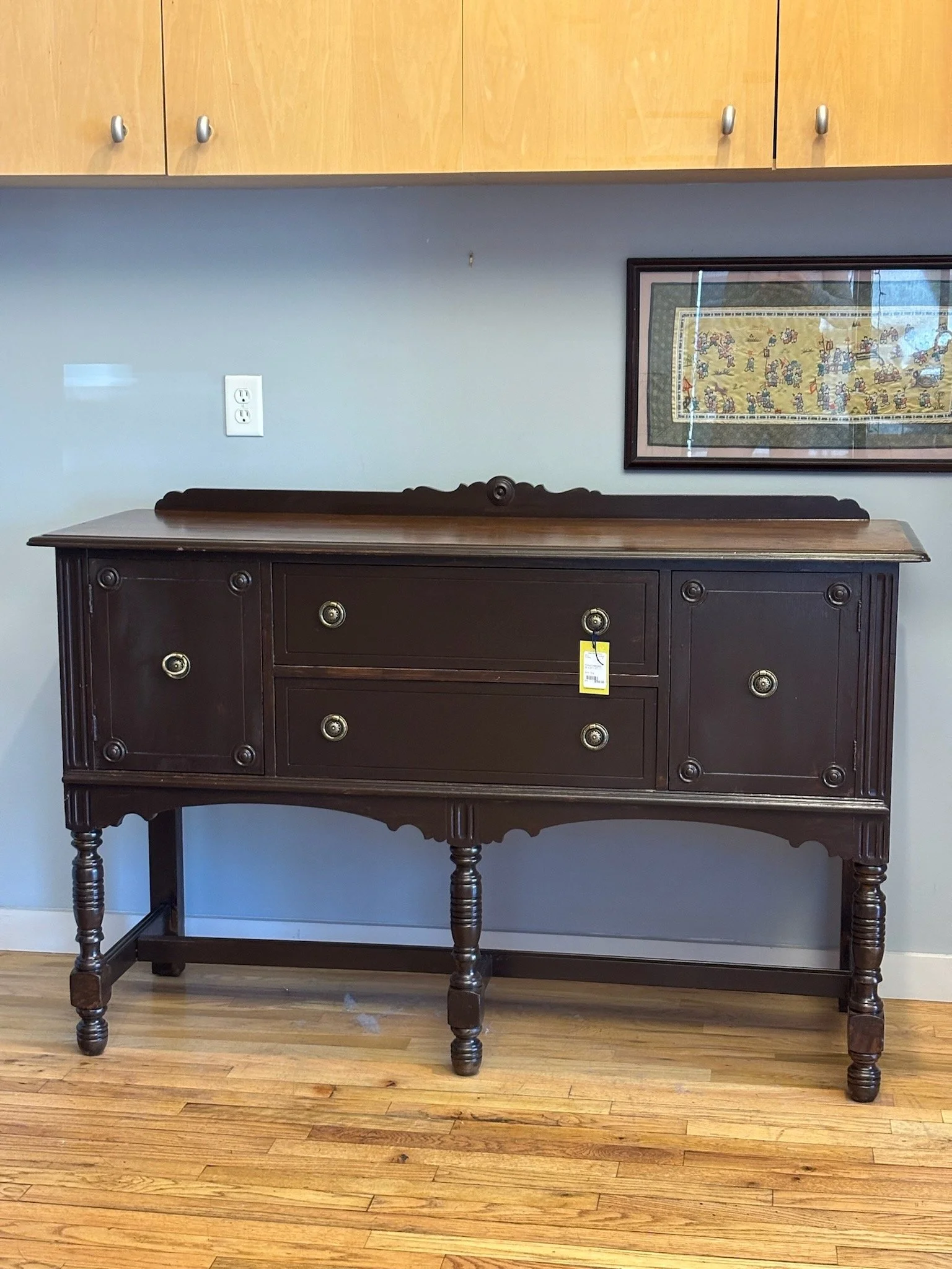 A dark wooden sideboard with four drawers and two cabinets, standing on four turned legs, against a gray wall with a framed colorful print and an electrical outlet, topped with a large wooden cabinet with four doors and silver knobs.