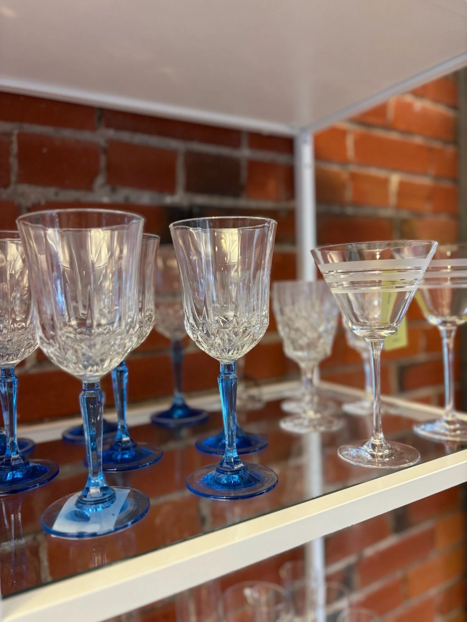 Collection of clear wine glasses with blue stems displayed on a glass shelf against a brick wall background.