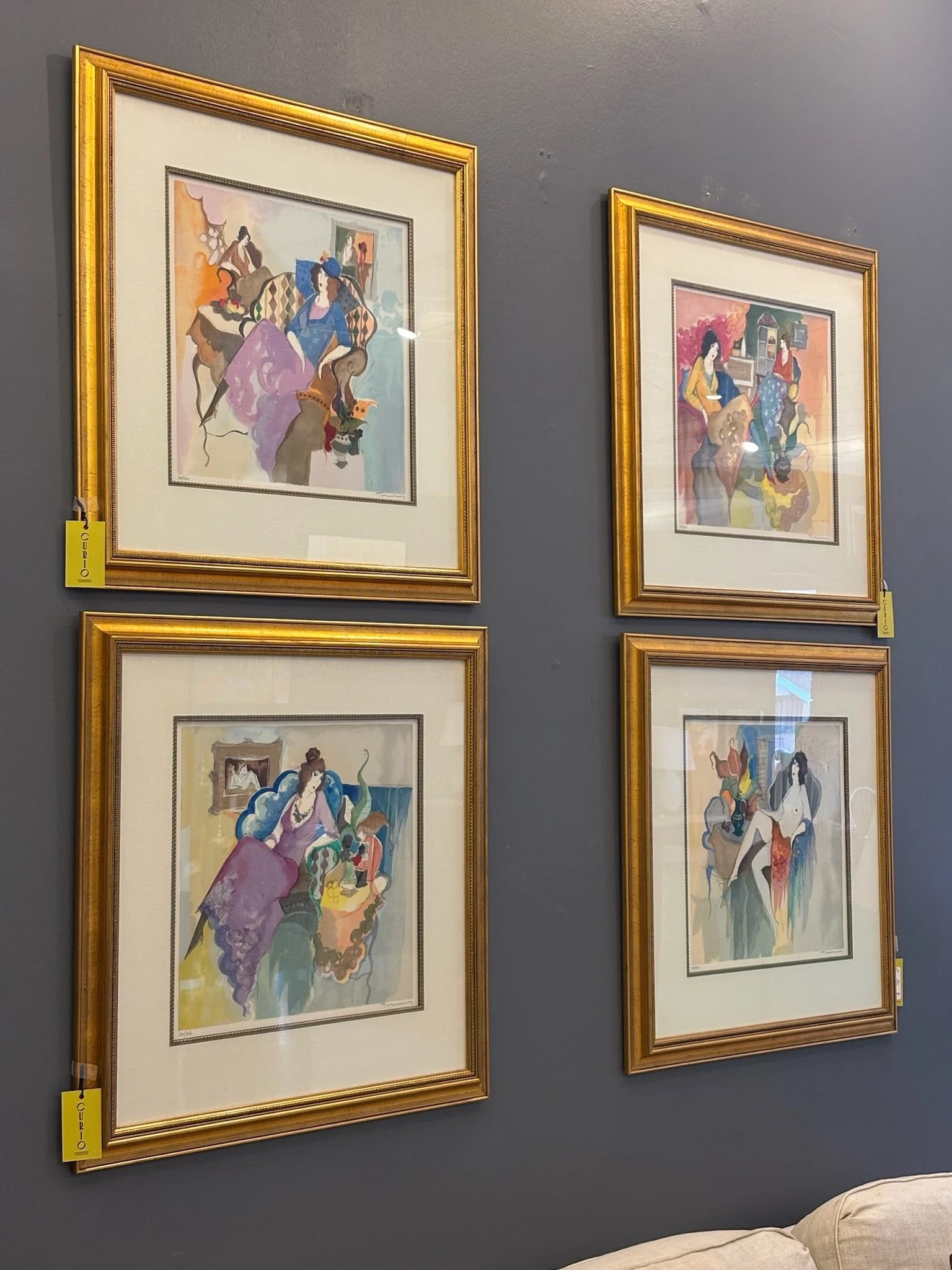 Four framed watercolor paintings of women in colorful dresses, hanging on a dark gray wall in an art gallery.