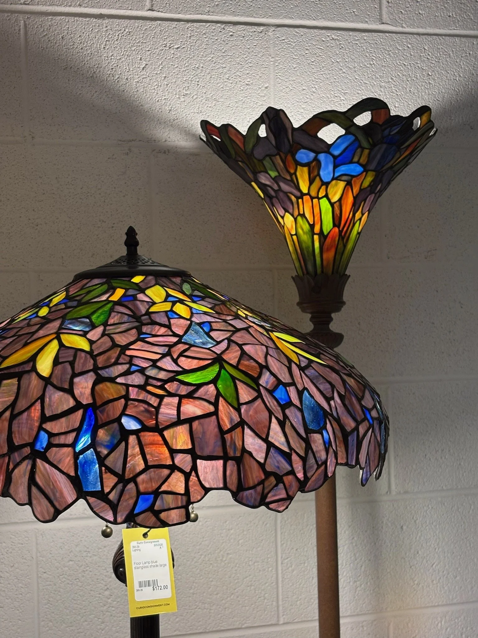 Colorful Tiffany-style floor lamp with a large stained glass shade featuring pink, purple, blue, yellow, and green glass pieces, on a wooden pole against a gray brick wall.