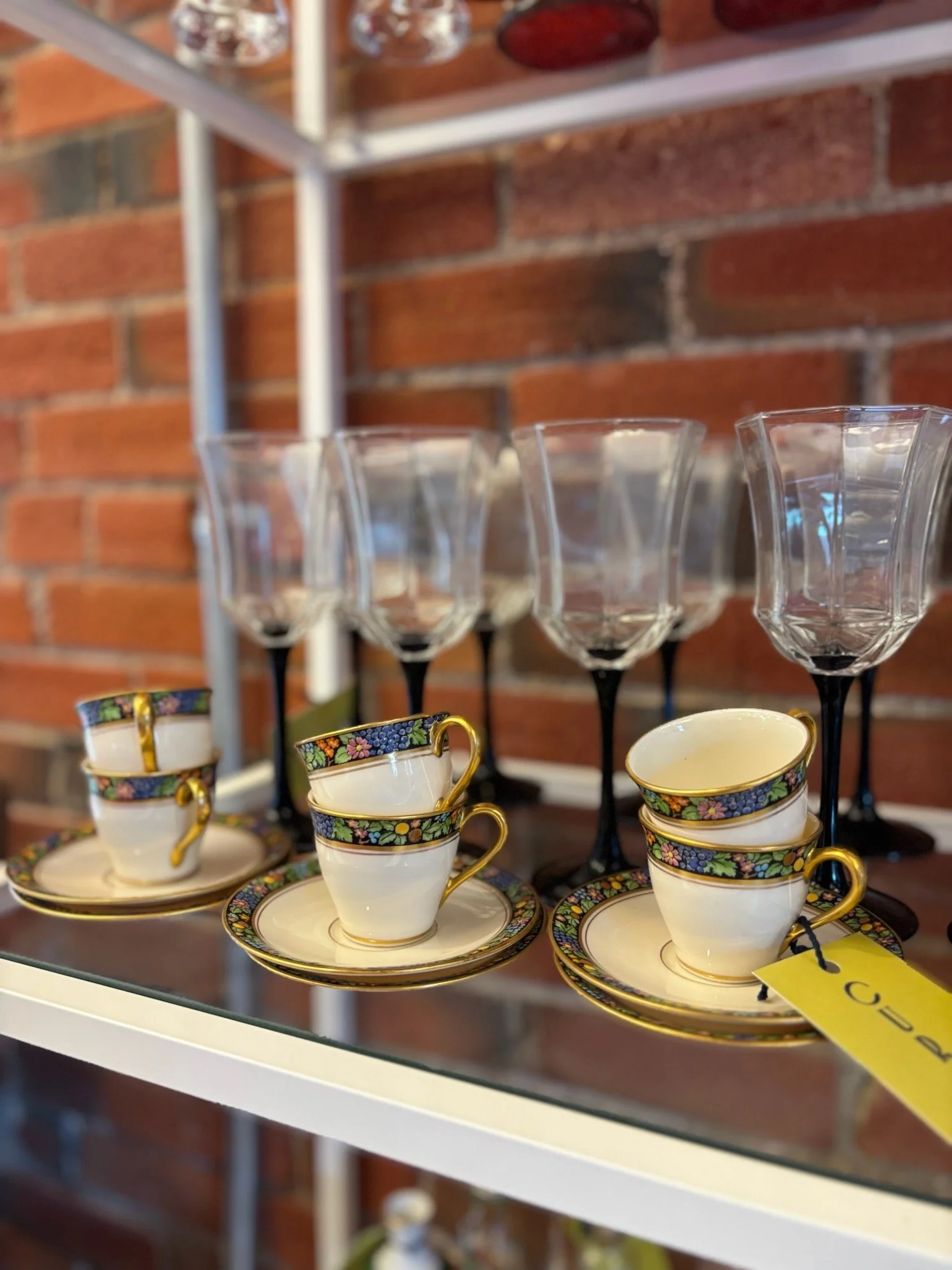 A display of elegant porcelain teacups with floral and gold detailing, arranged on matching saucers, with crystal glasses and shelves with decorative items in the background.